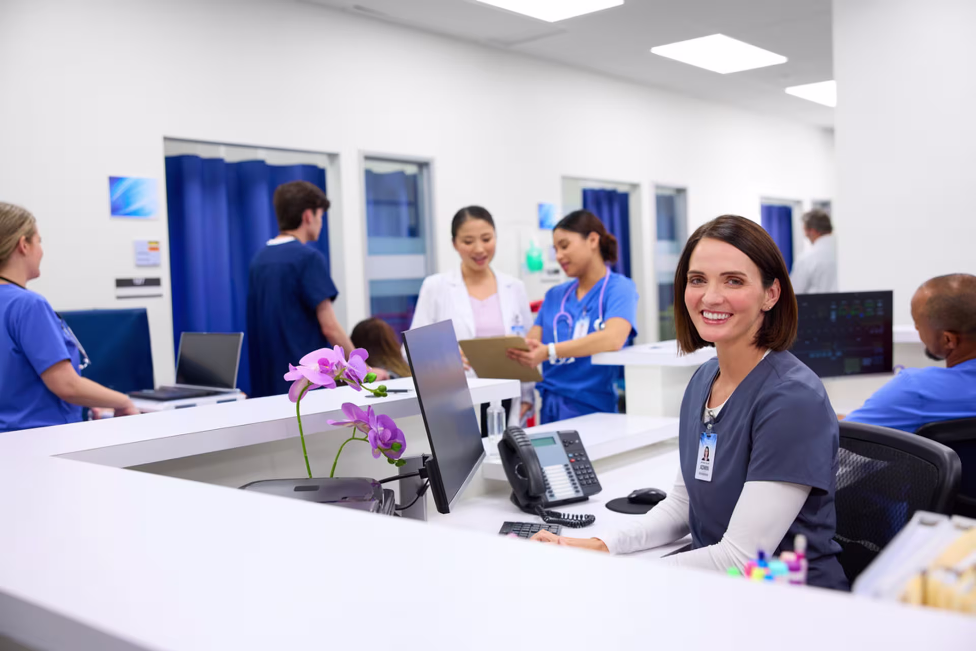 Smiling healthcare administrator at hospital reception desk with nurses and medical staff collaborating in modern clinic lobby.