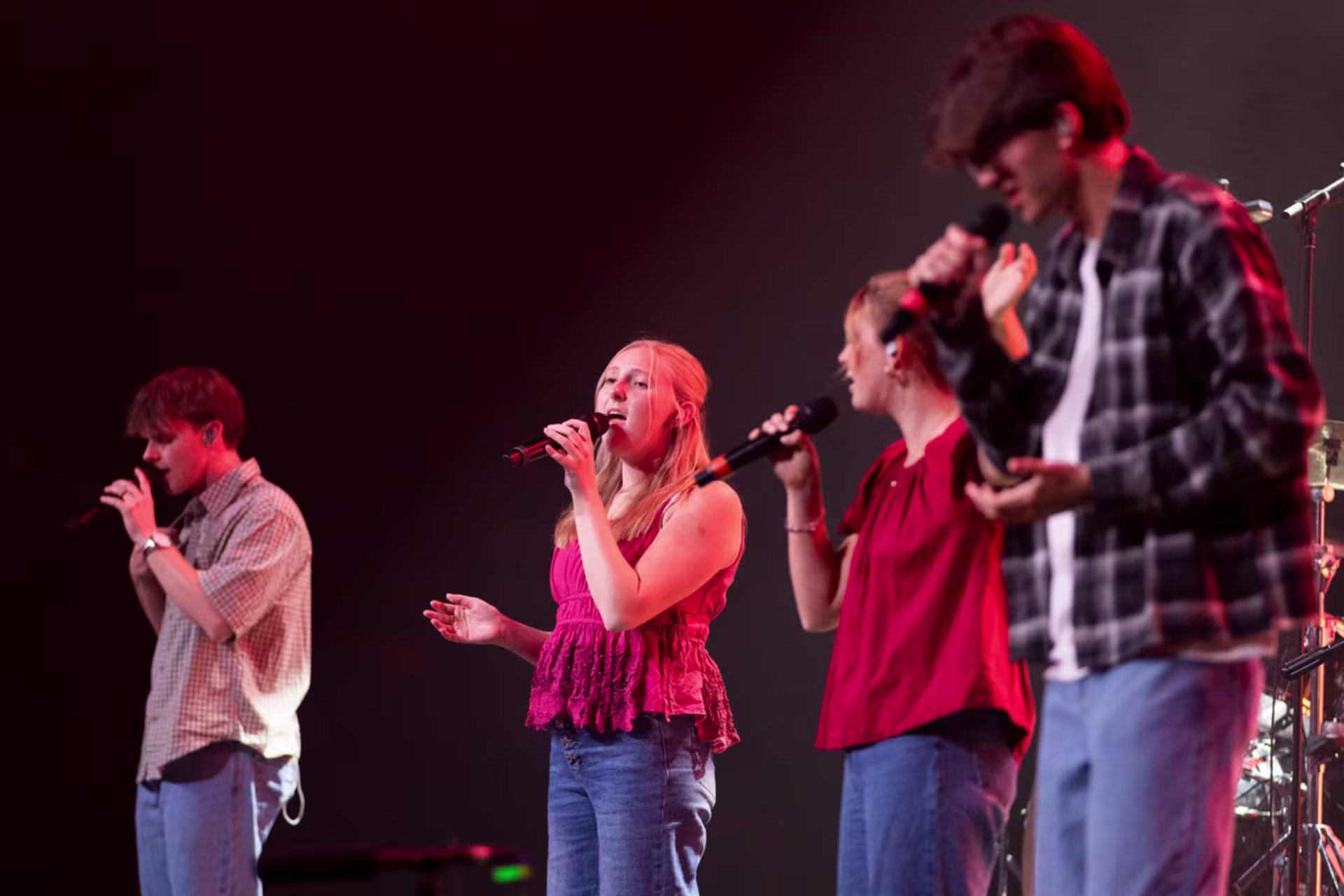 A group of singers performs on stage with handheld microphones under red lighting.