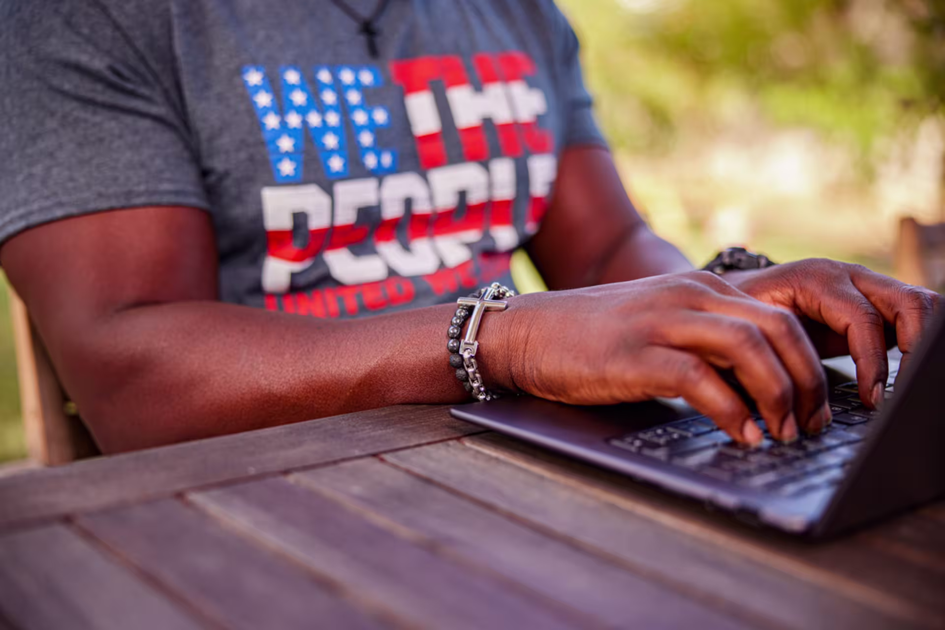 Male typing on a laptop at an outdoor wooden table, with a casual T-shirt and blurred greenery in the background.