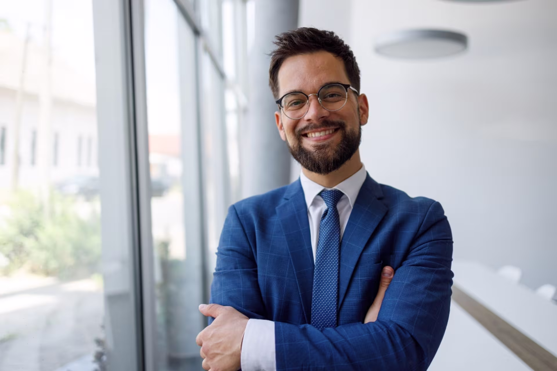 A business professional smiles in an office hallway with his arms crossed.