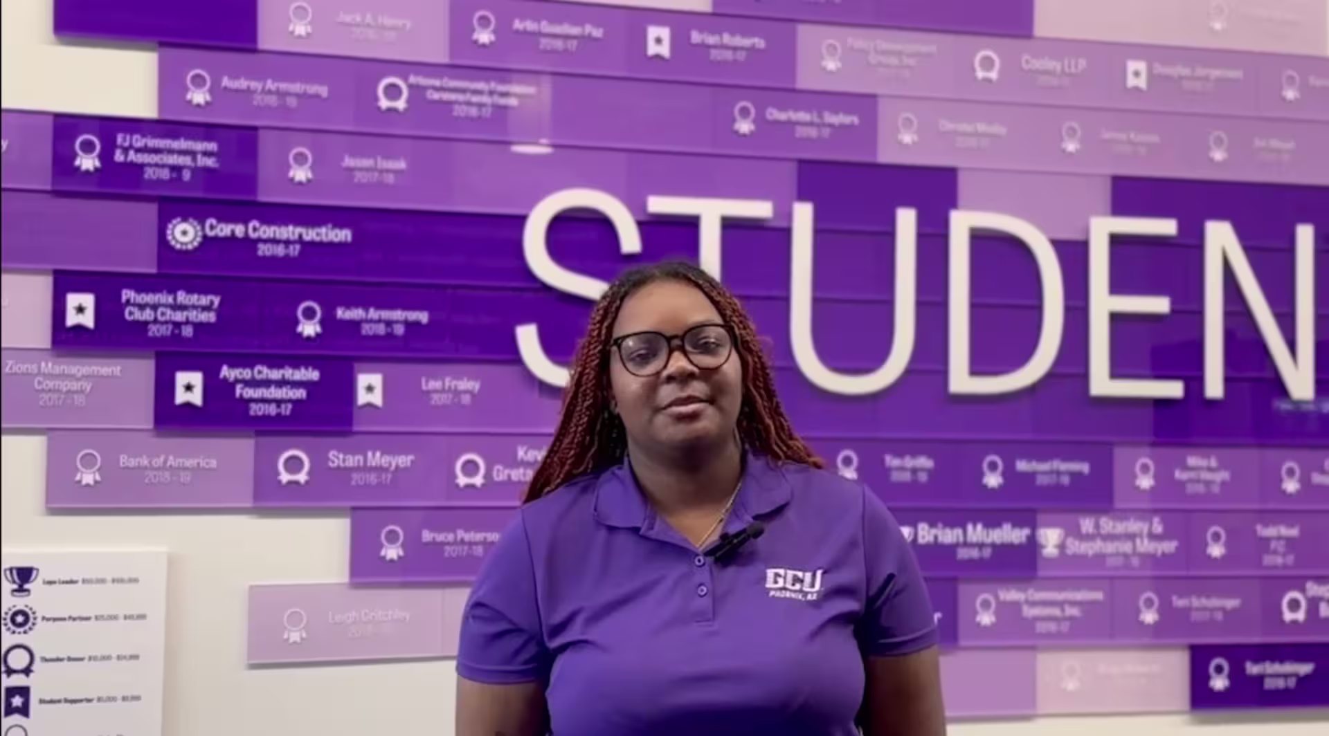 Female wearing a purple GCU polo standing in front of a purple wall display with “STUDENT” text and listed program categories in the background.