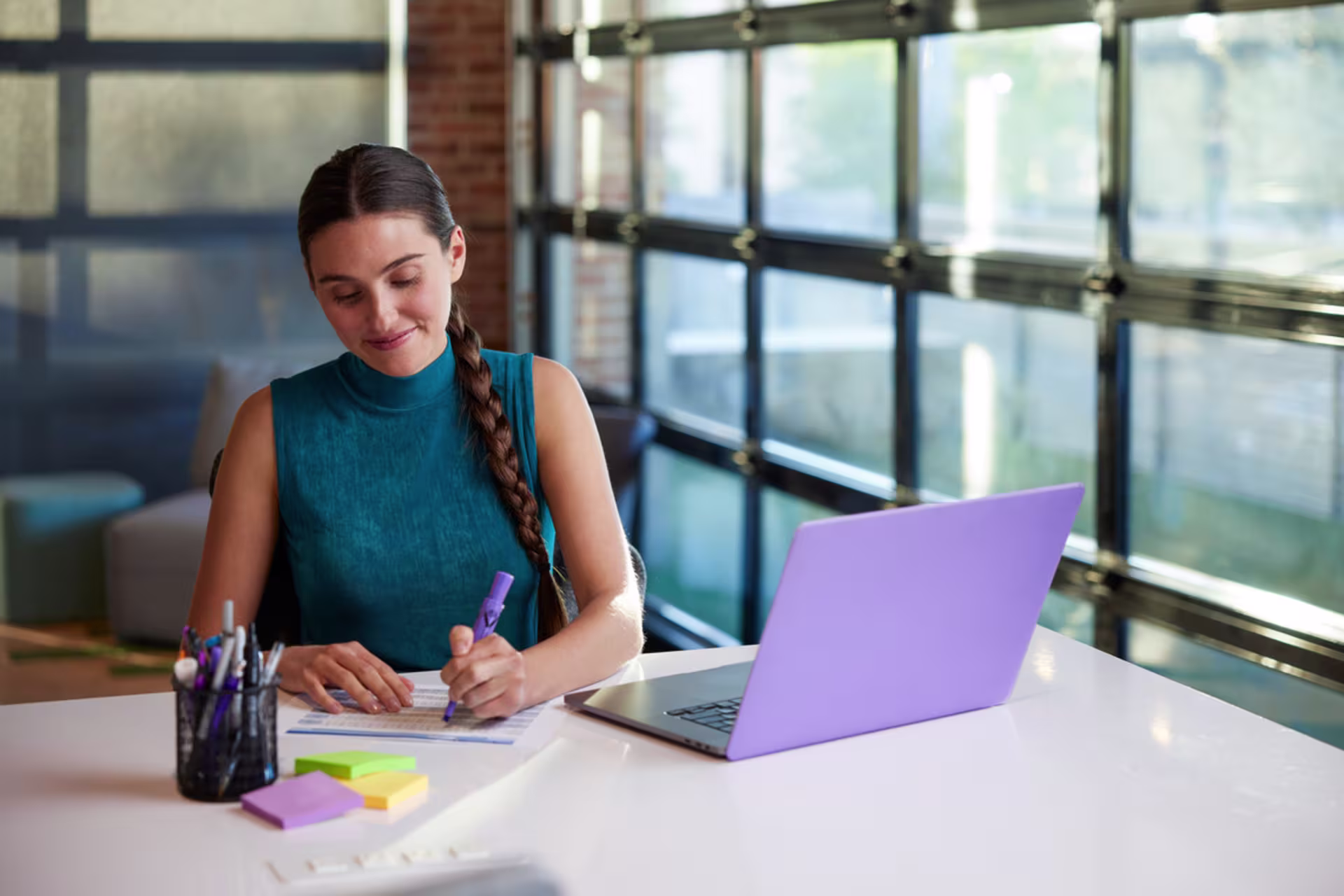 A female copywriter sits at a desk by large windows, writing in a notebook with a pen, with a purple laptop open beside her, along with a cup holding pens and a stack of colorful sticky notes.