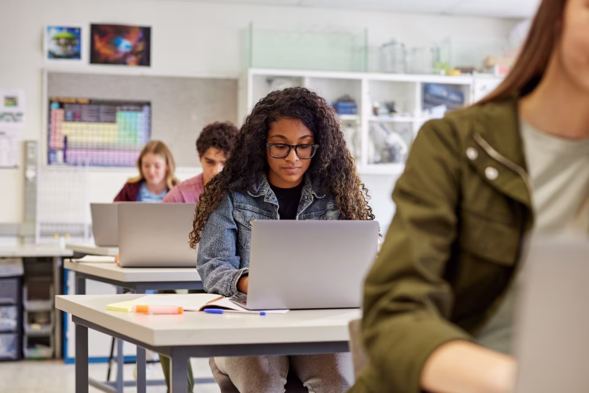 A high school student with curly hair and glasses sits at a classroom desk focused on a laptop, with notebooks and pens spread out in front of her.