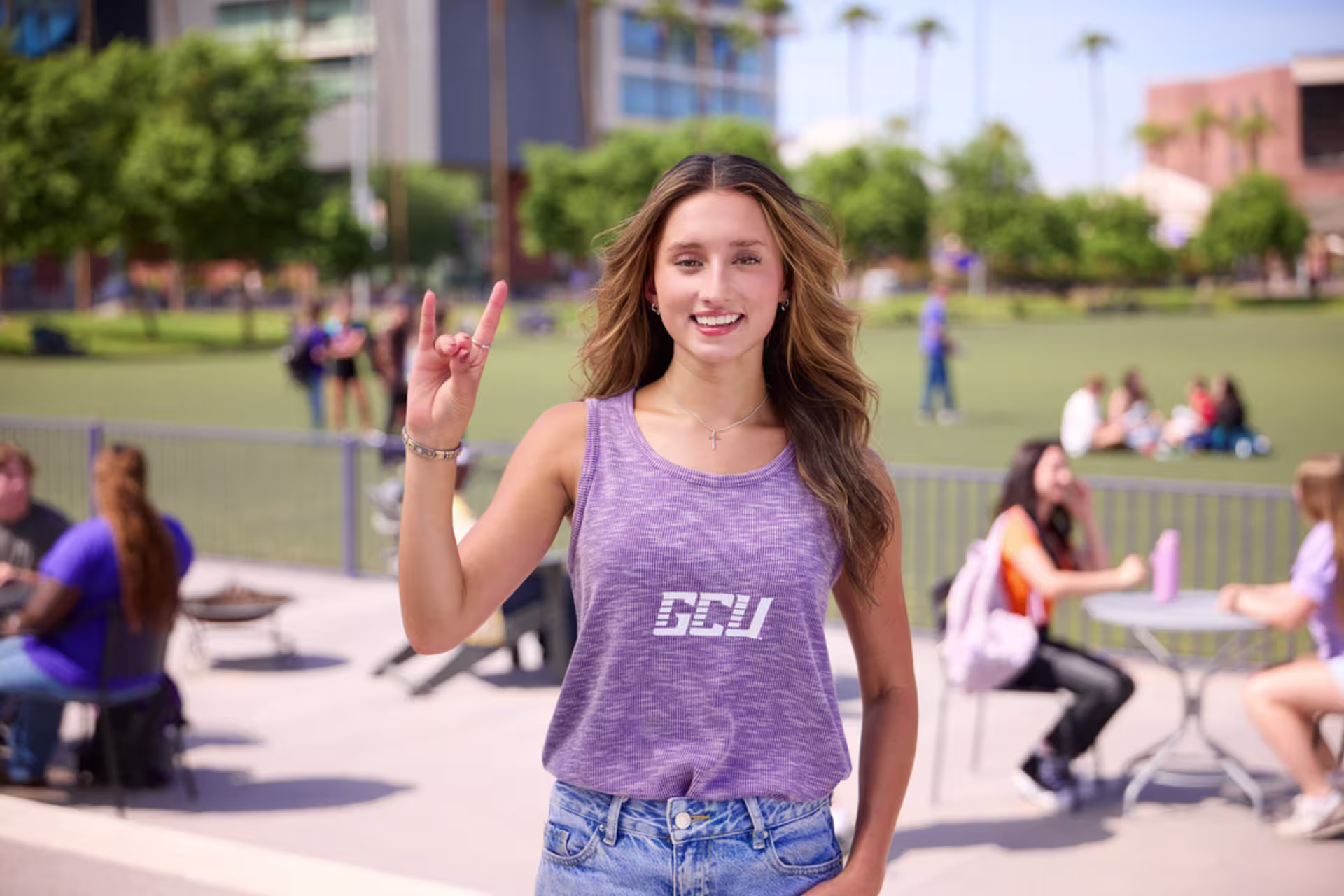 A female GCU student in a purple tank top stands on a campus lawn raising the Lopes Up sign with students and university buildings in the background.