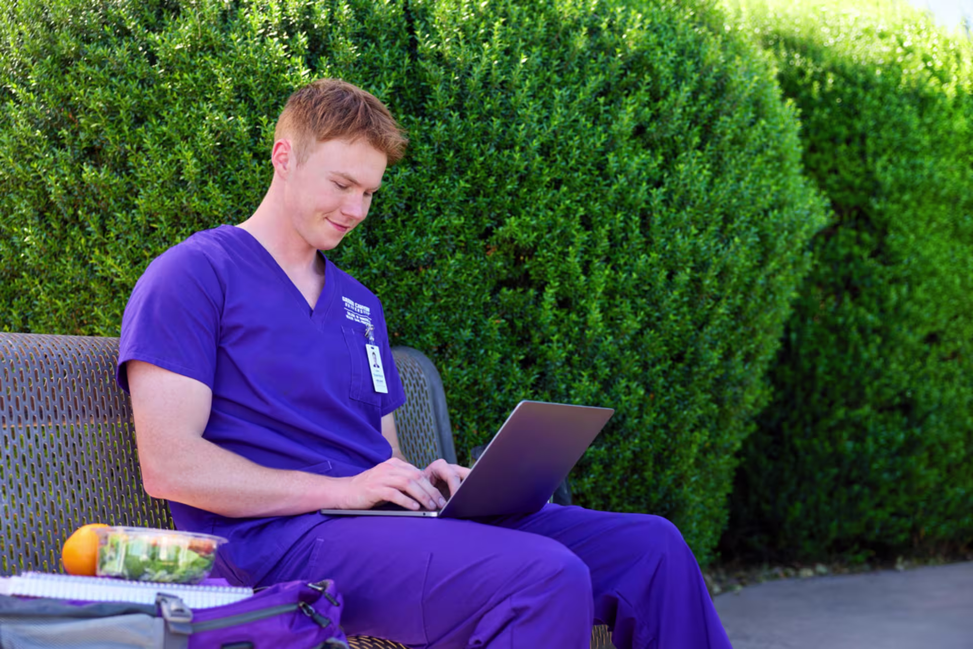 Male GCU nursing student wearing purple scrubs sitting on bench with open laptop doing online coursework.