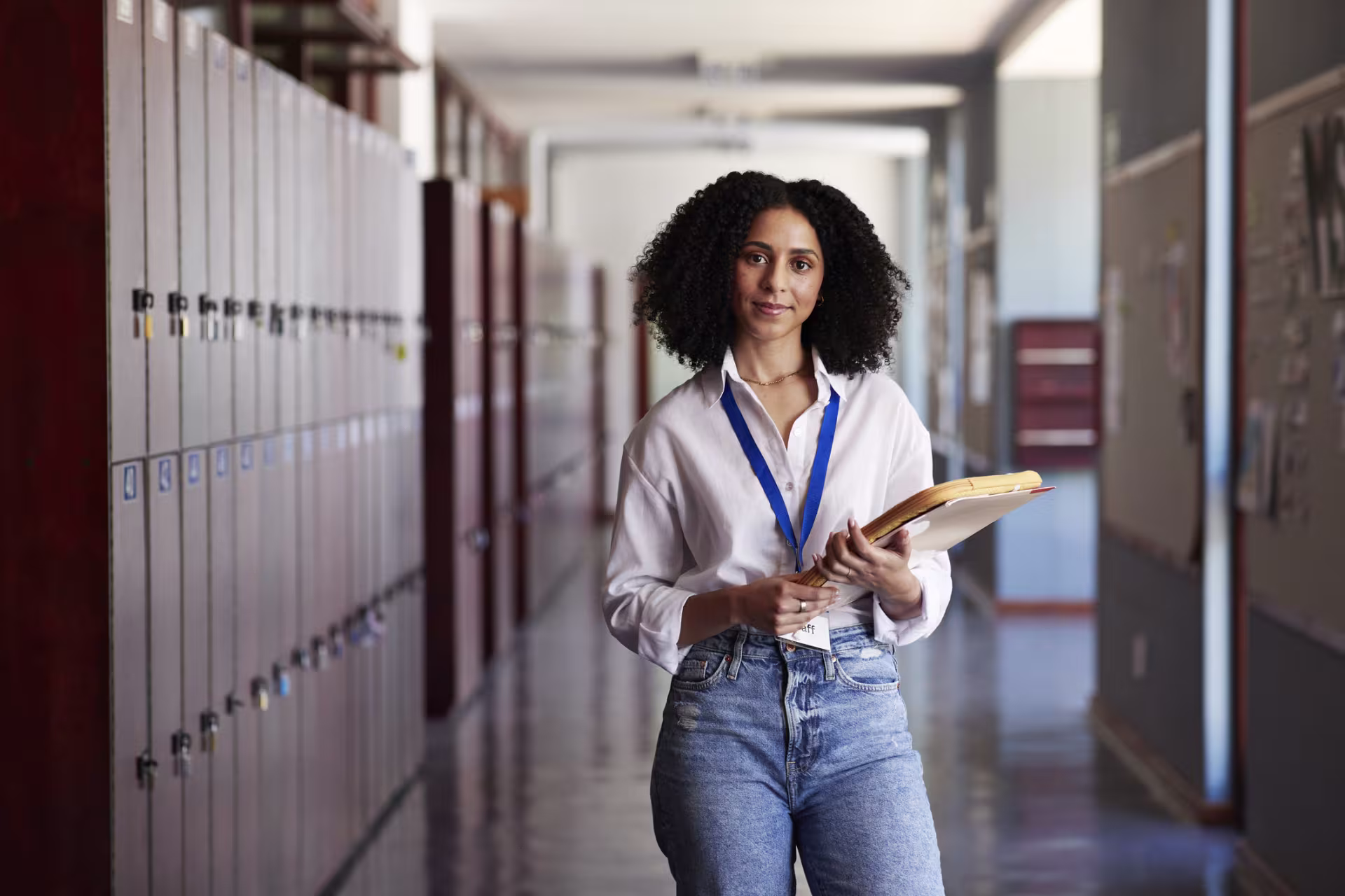 Educator wearing a lanyard walks through a high school hallway while holding academic materials.