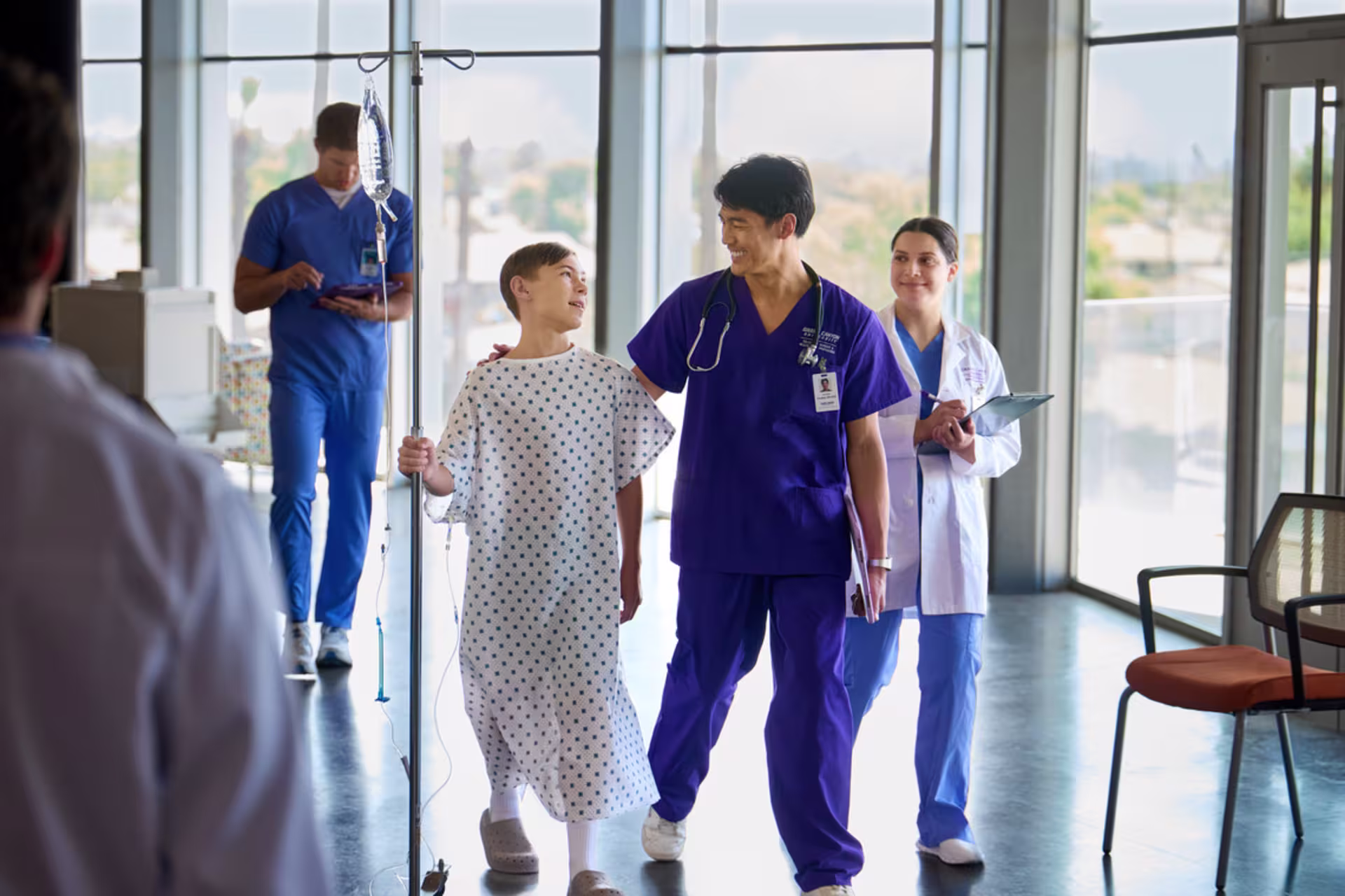 A male GCU nursing student in purple scrubs walks with a patient during hospital clinical training.