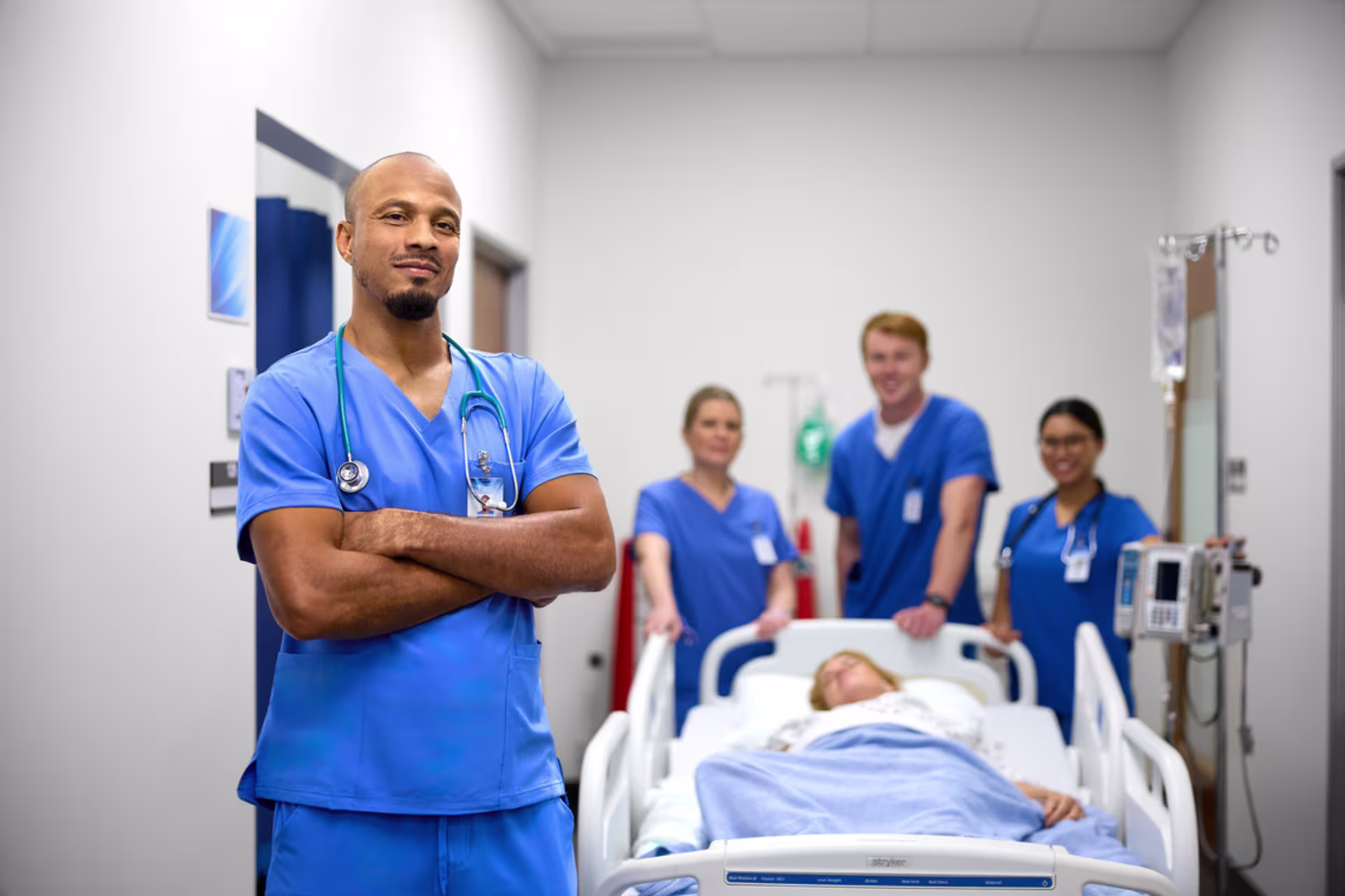 A nursing team in blue scrubs stands confidently in a clinical setting, with a patient resting in a hospital bed in the background.
