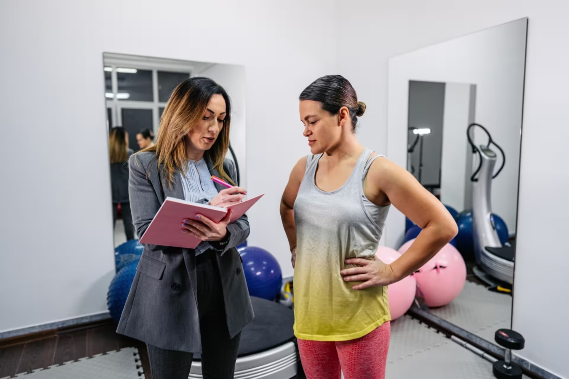 Female sport and performance psychologist writes notes on a clipboard while meeting with a patient in gym attire in an exercise room.