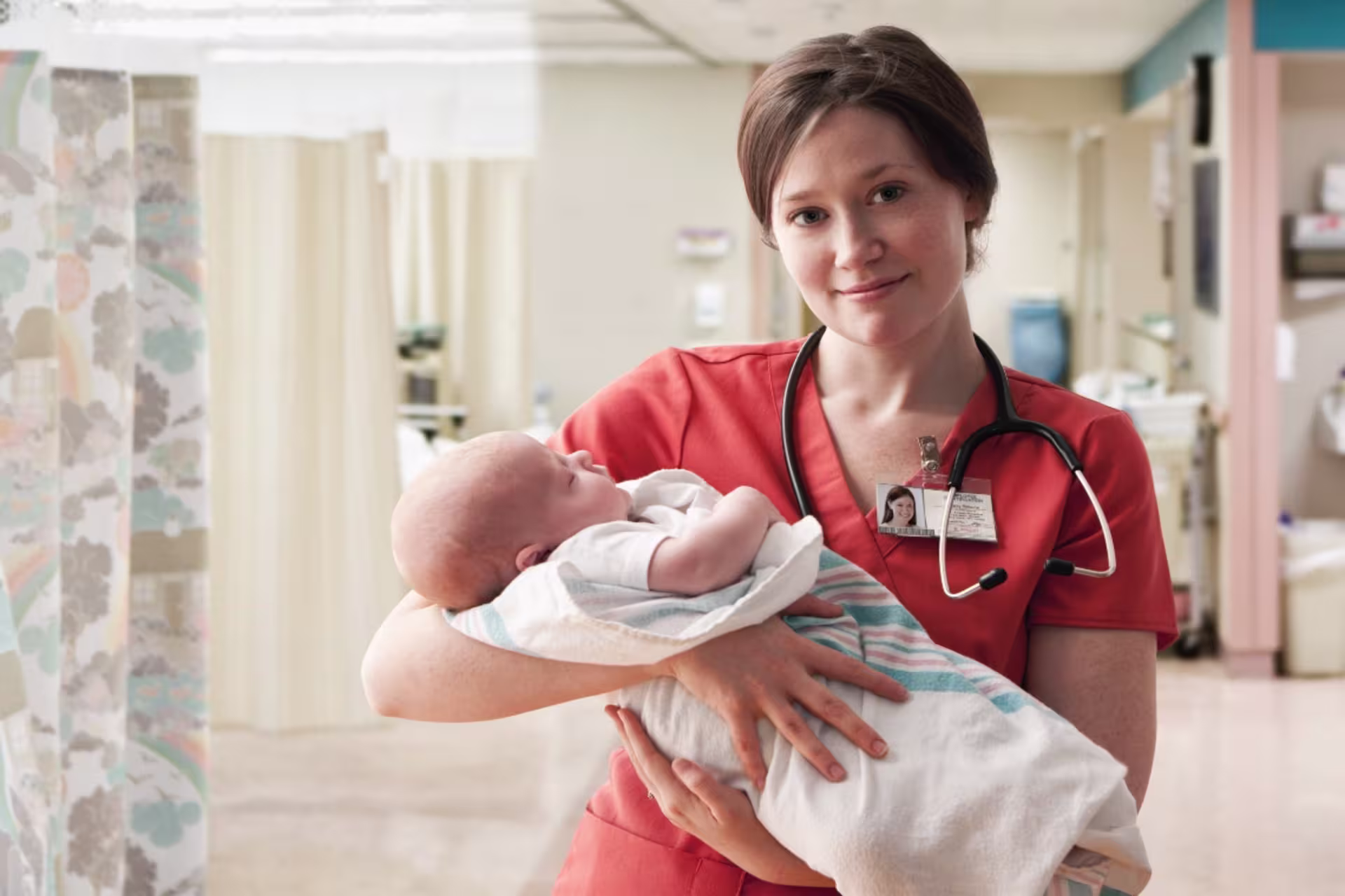 A female labor and delivery nurse holds a newborn baby in a hospital setting.