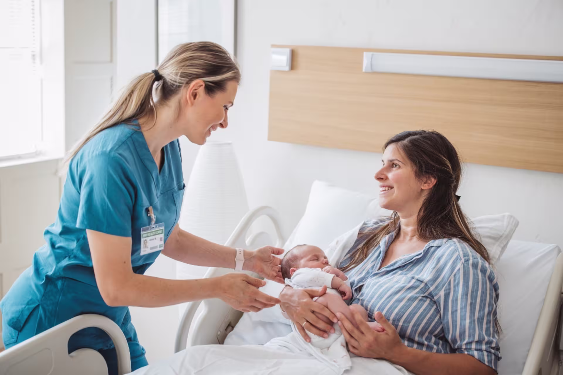 Nurse helping a new mother hold her newborn baby in a hospital maternity room.