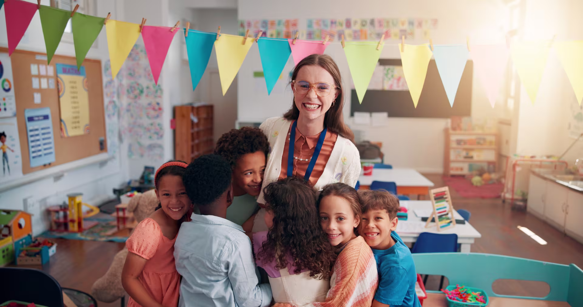 A teacher stands in a colorful classroom while several young children gather closely around in a group hug, with bright pennant banners hanging overhead and classroom materials visible in the background.