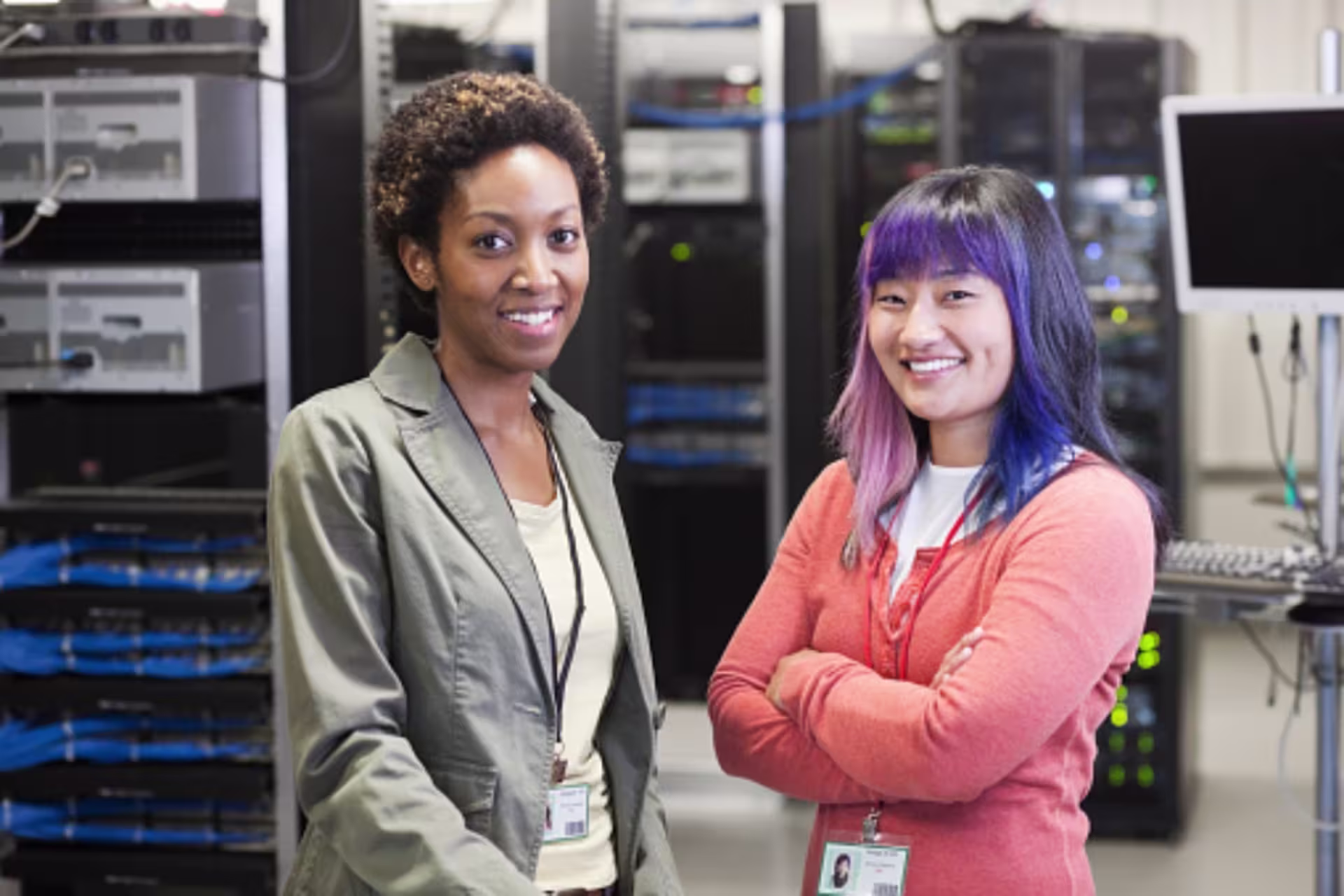 Two female students wearing ID badges stand in a server room, with rows of server racks, cables and computing equipment visible behind them.