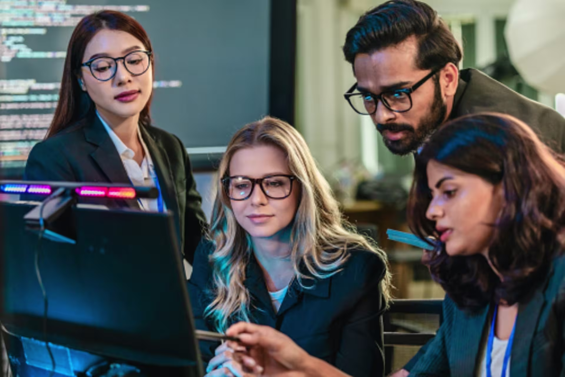 A group of people gathered around a computer work together while looking at a screen in a professional office or tech workspace.