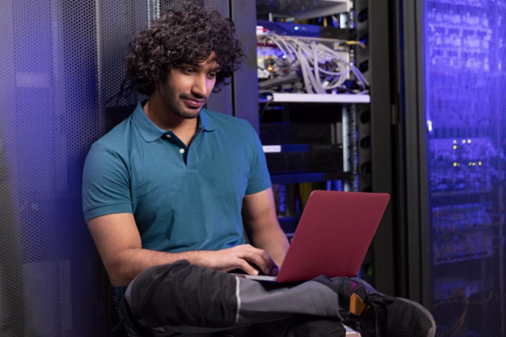 A network administrator sits in a server room, using a laptop beside rows of network and computer equipment.