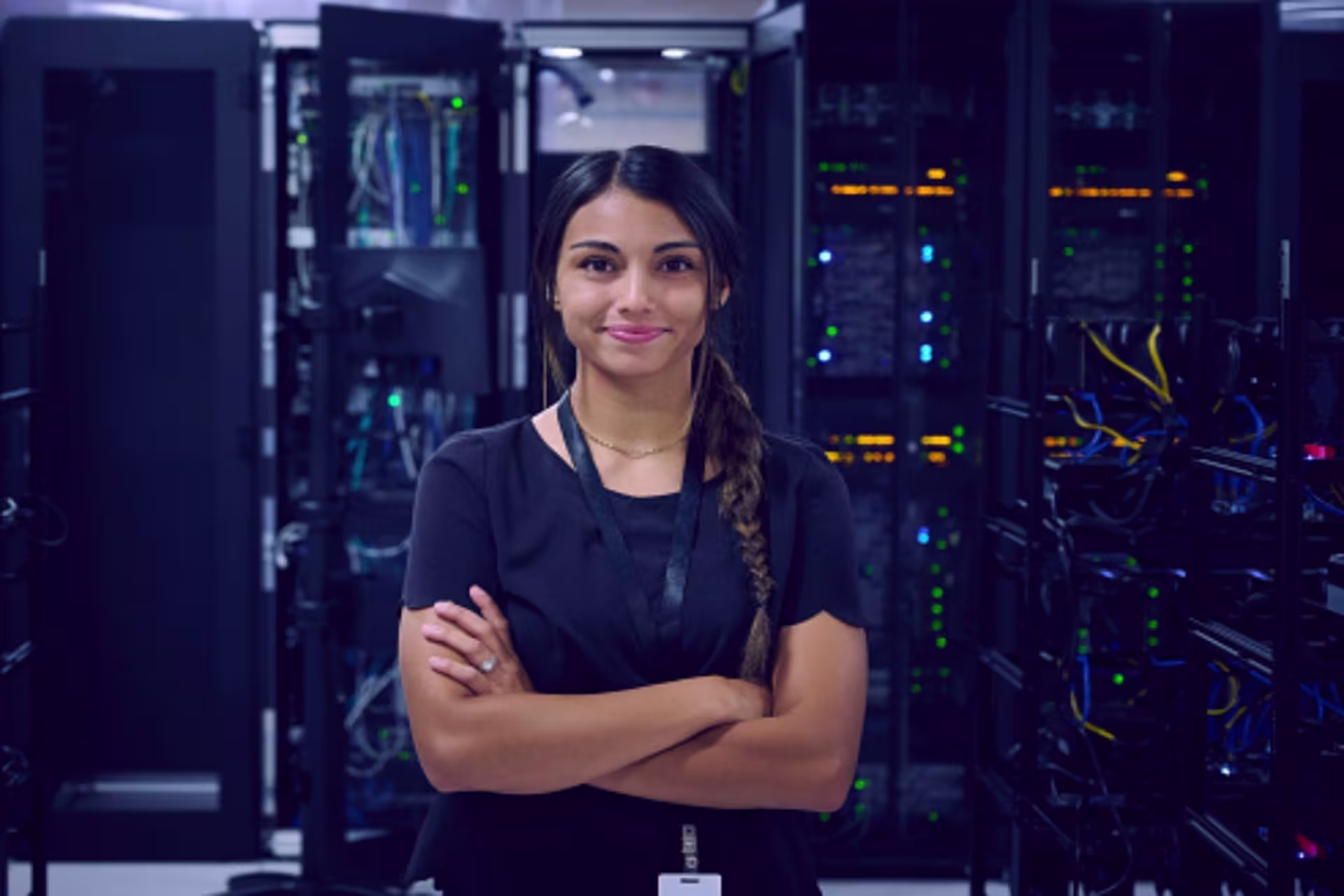 A female networking administrator stands with arms crossed in a server room, with rows of computer servers and cables in the background.