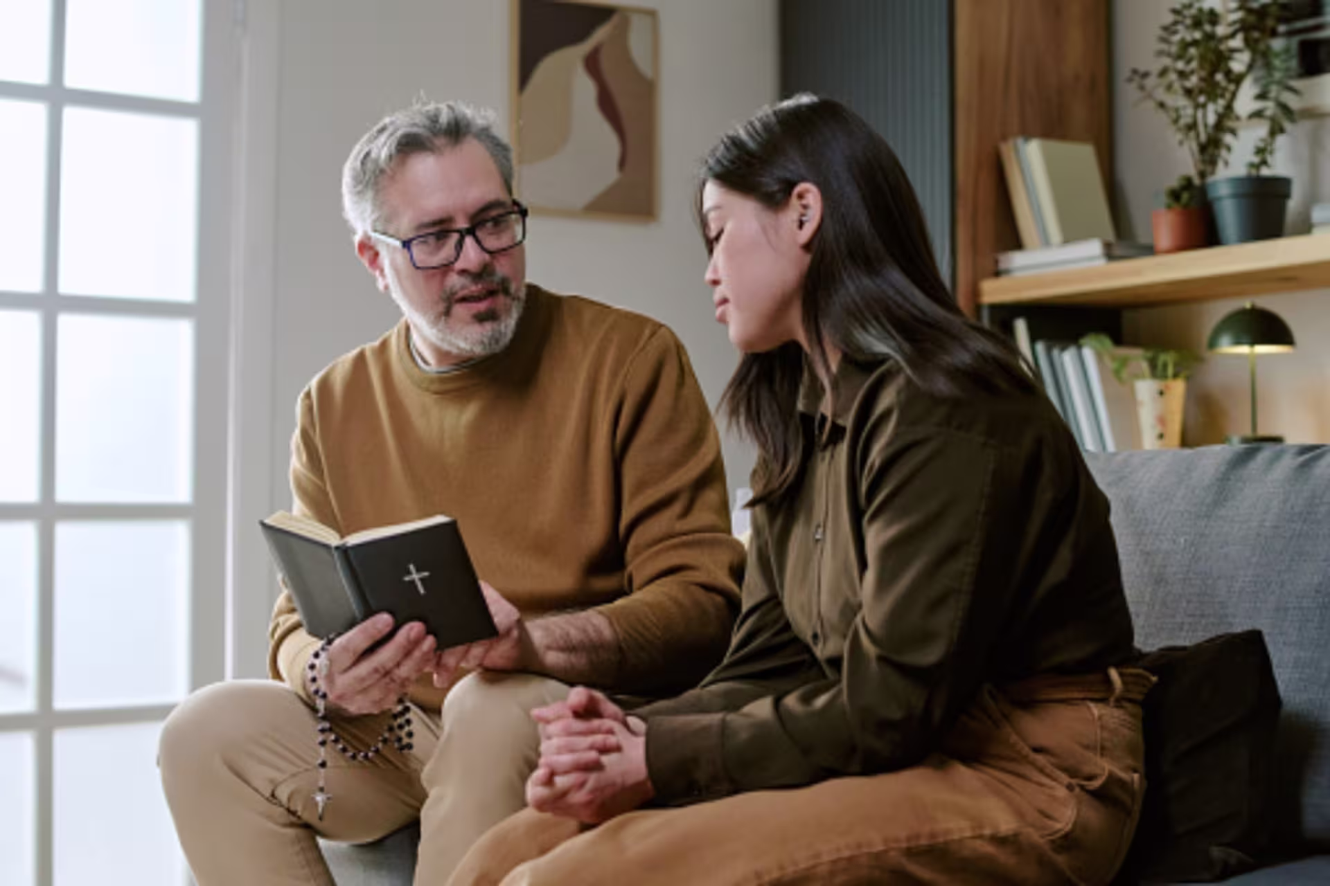 A pastor holds a Bible and rosary while speaking with a young woman during a counseling session in a home setting.