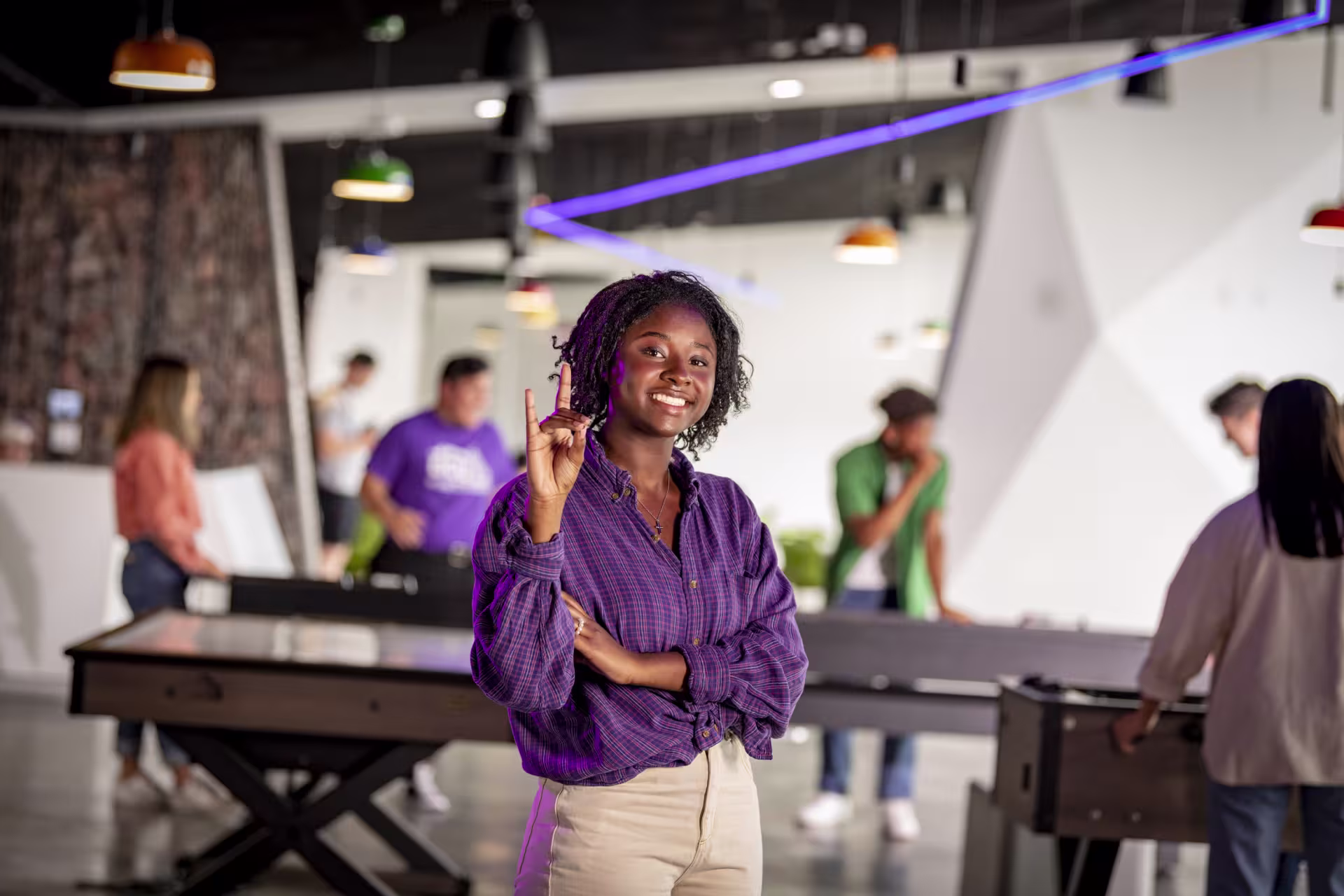 Lela Lewis stands in a student recreation space with pool tables, posing with one hand raised while looking toward the camera, with other people and colorful accent lighting in the background.