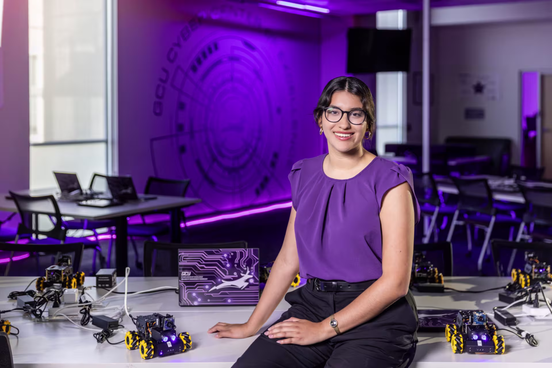A female GCU cybersecurity student poses in a technology lab with robotics equipment.