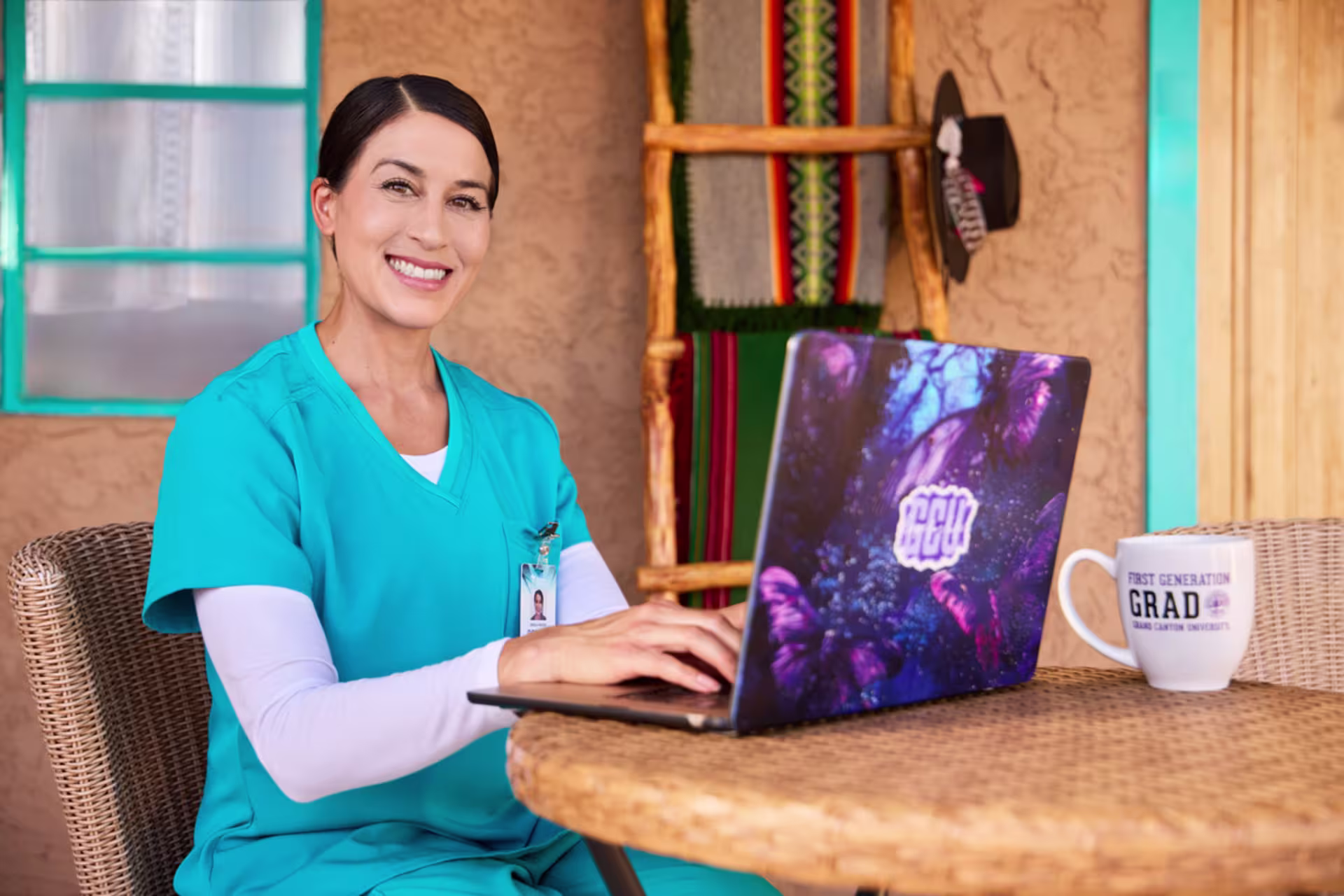 A smiling nursing student studies online, using a laptop outside on a back patio near an adobe home.