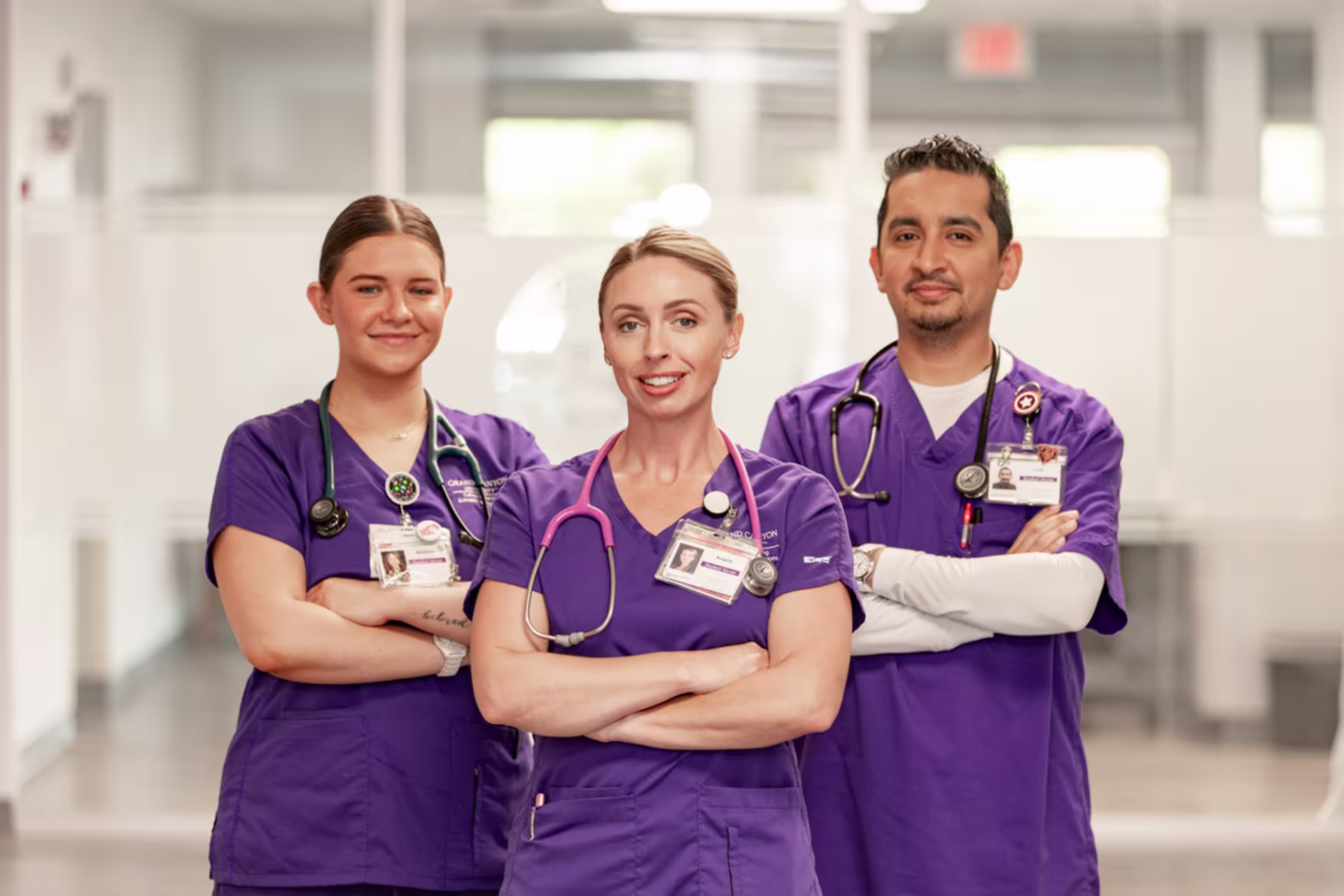 Nursing degree students in scrubs stand together with their arms crossed while looking straight ahead in a hospital setting.