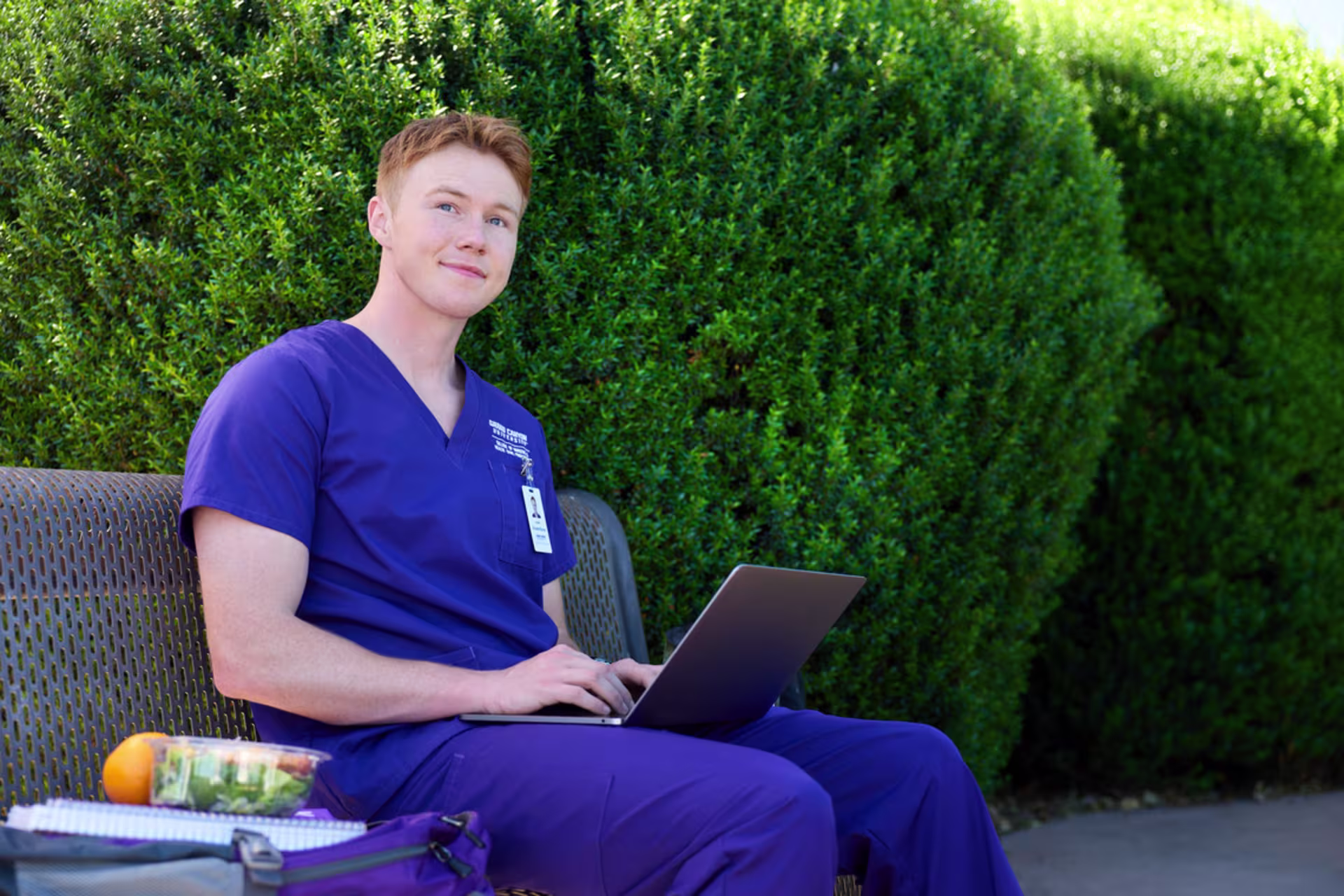 A male nursing degree student in purple scrubs uses a laptop outdoors.
