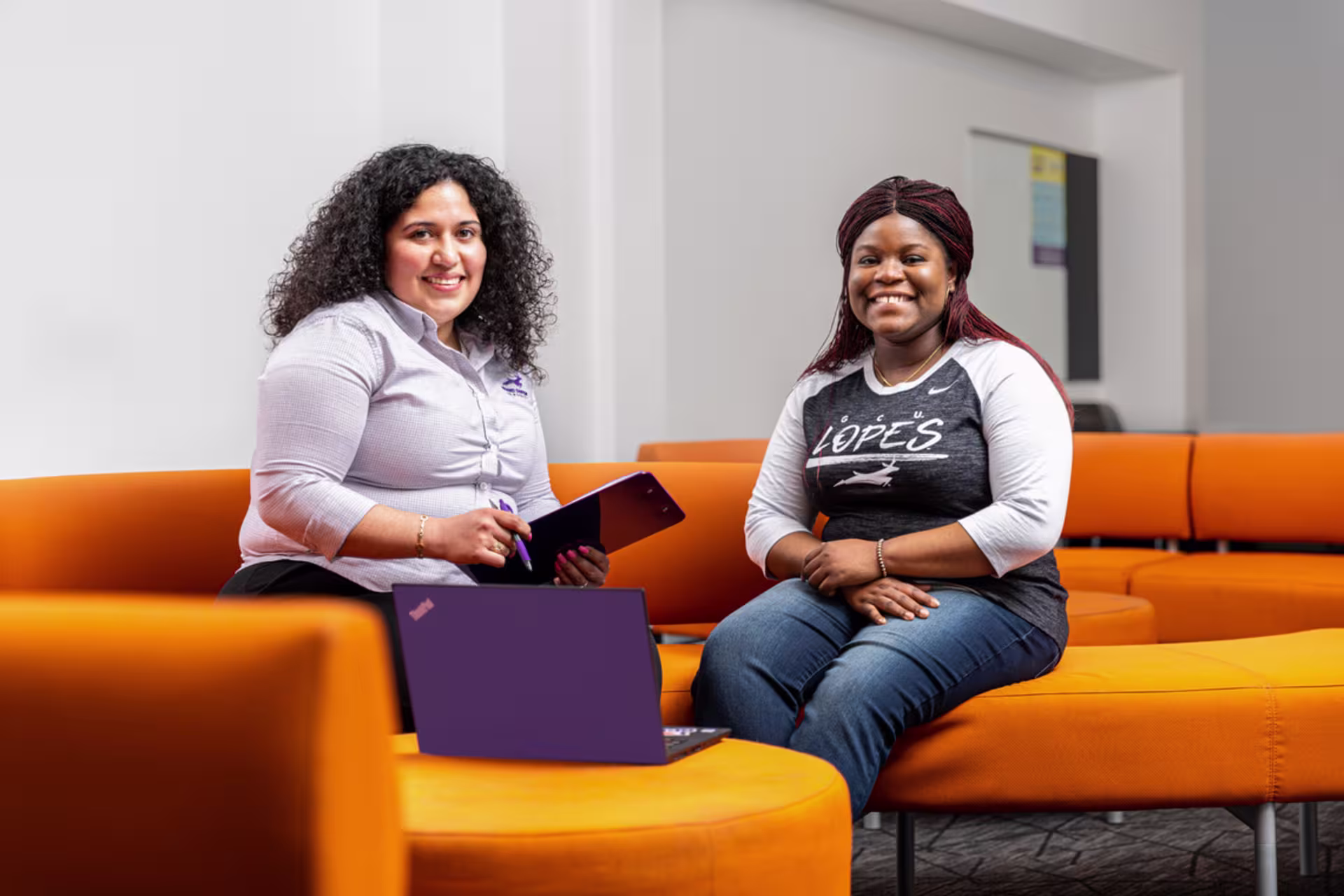 Two nursing students smile while seated on an orange couch in a bright campus lounge.