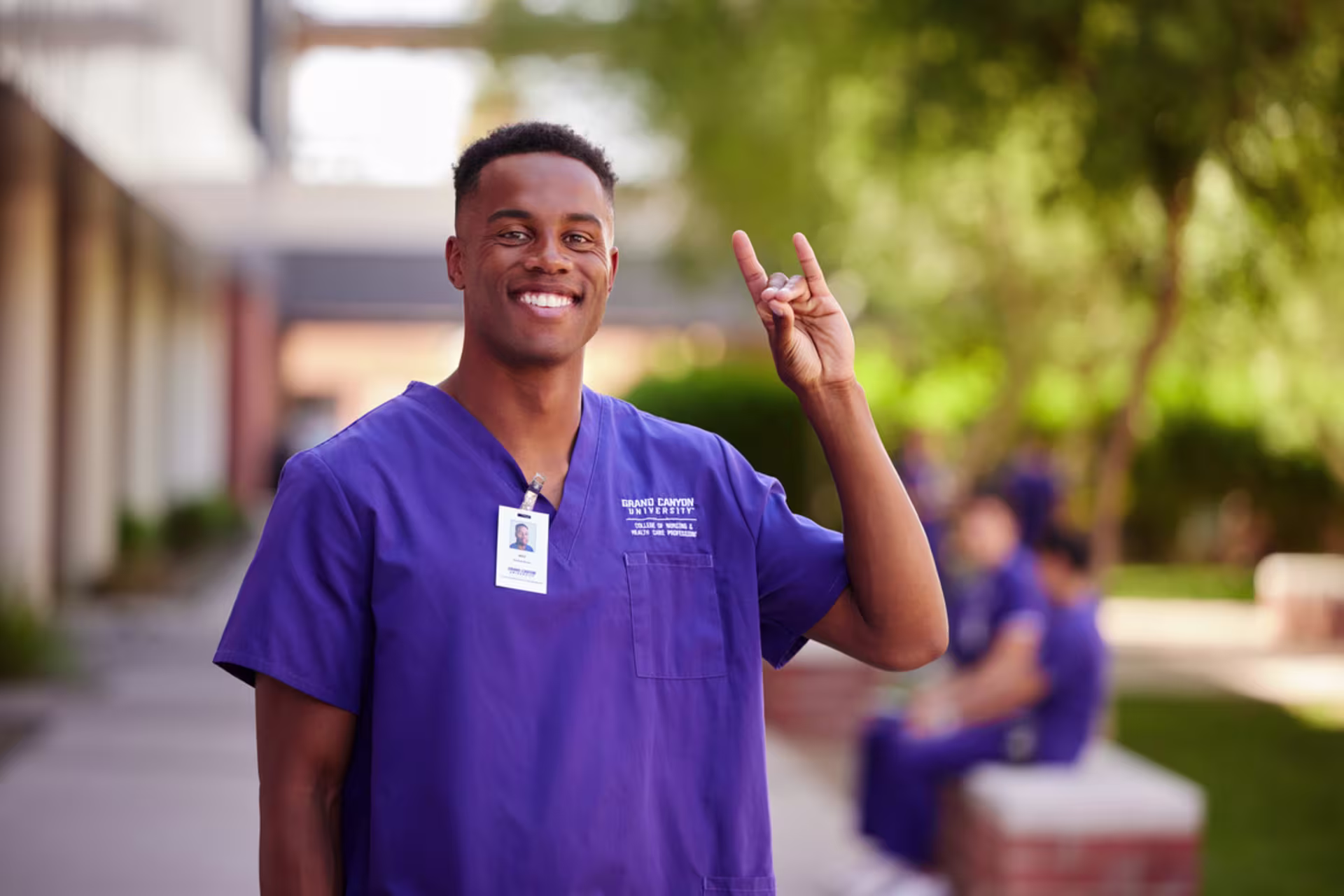 A male nursing student in scrubs poses on campus, smiling while holding the Lopes Up sign.