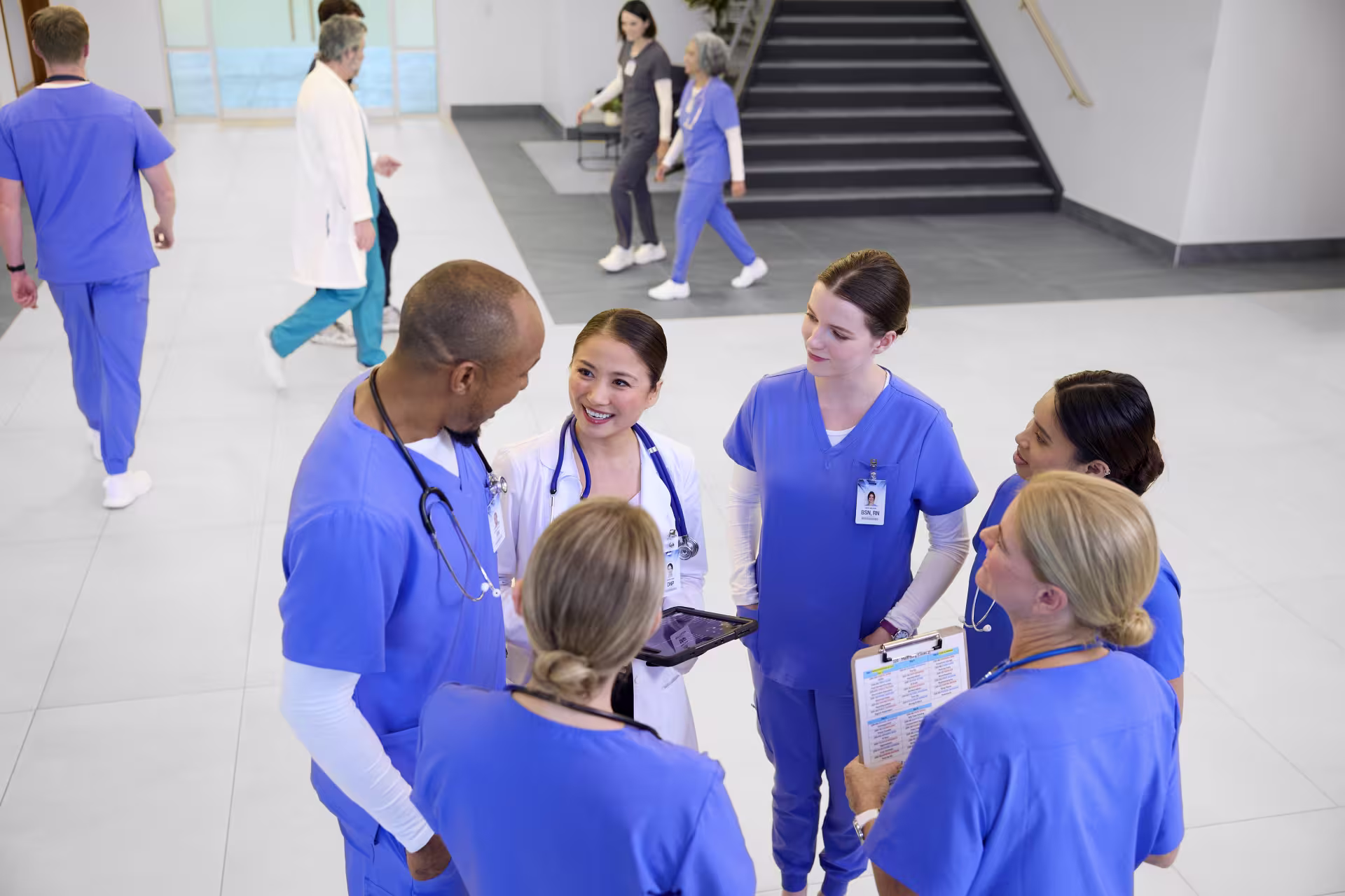 A group of healthcare professionals in blue scrubs and a white coat gather in a hospital lobby for a team discussion while other staff walk through the background.