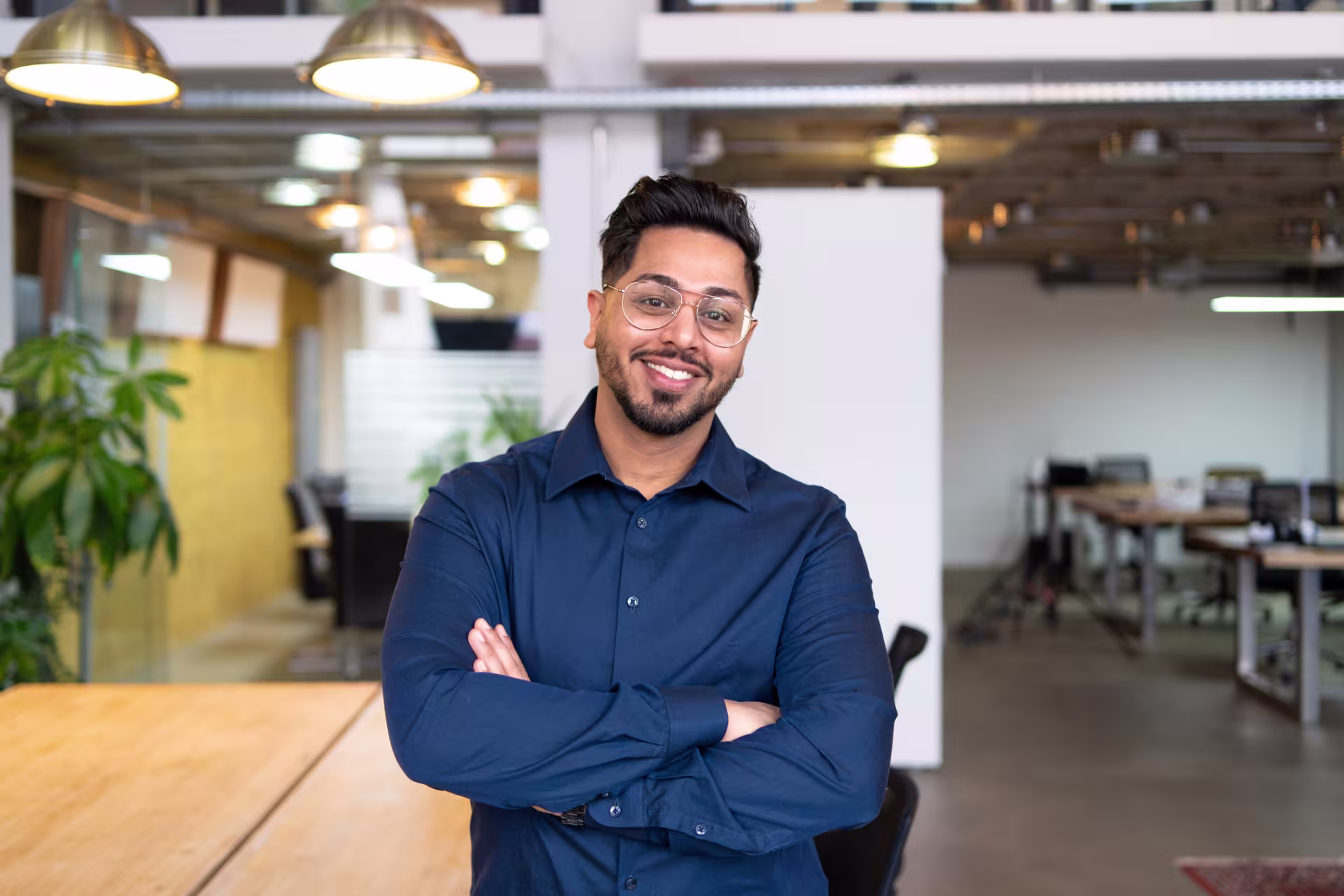Portrait of a smiling male wearing a navy button-down shirt and glasses, standing with arms crossed in a bright, open-plan office.