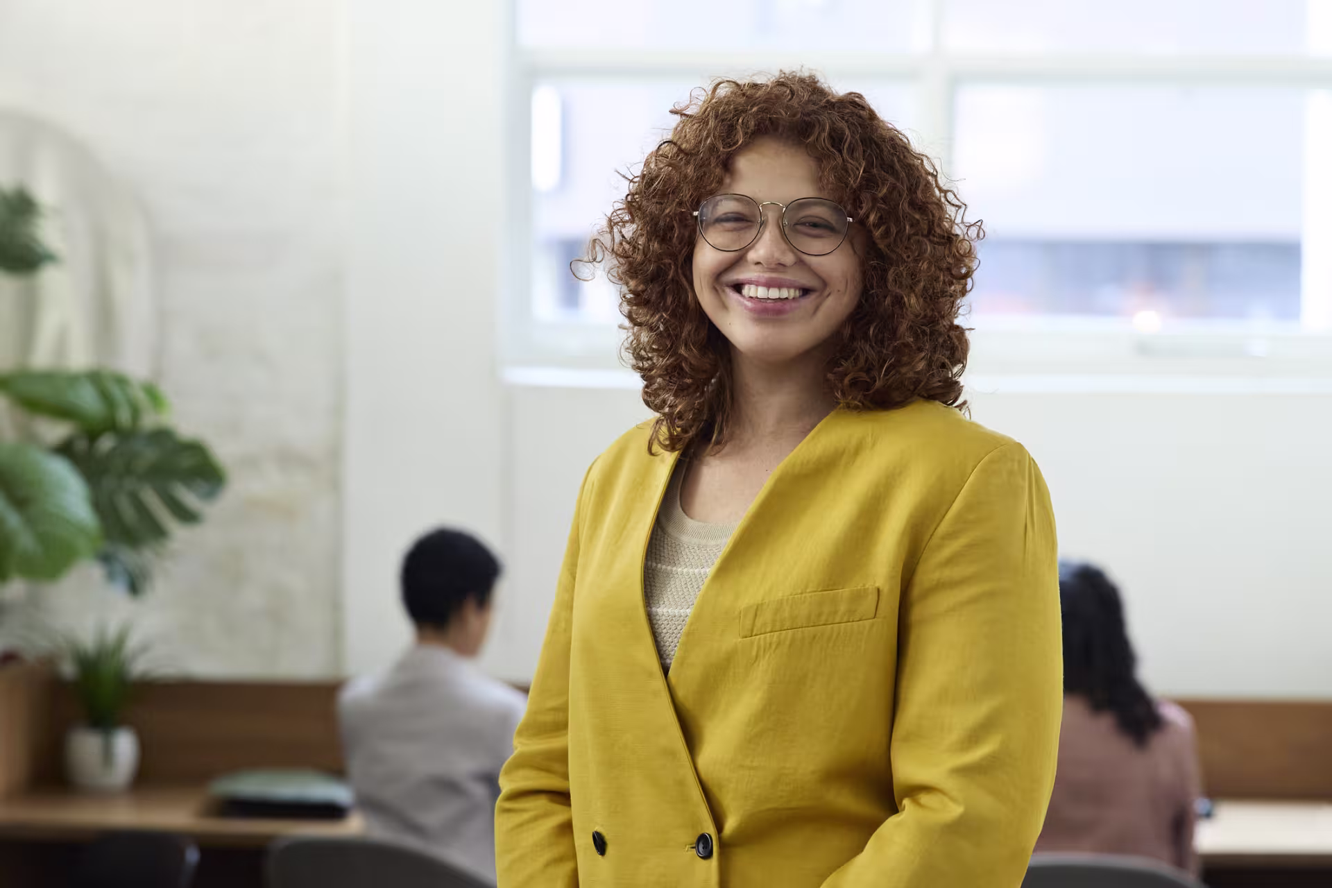 Confident, smiling woman wearing a yellow blazer and glasses in a bright office or study setting with others in the background.