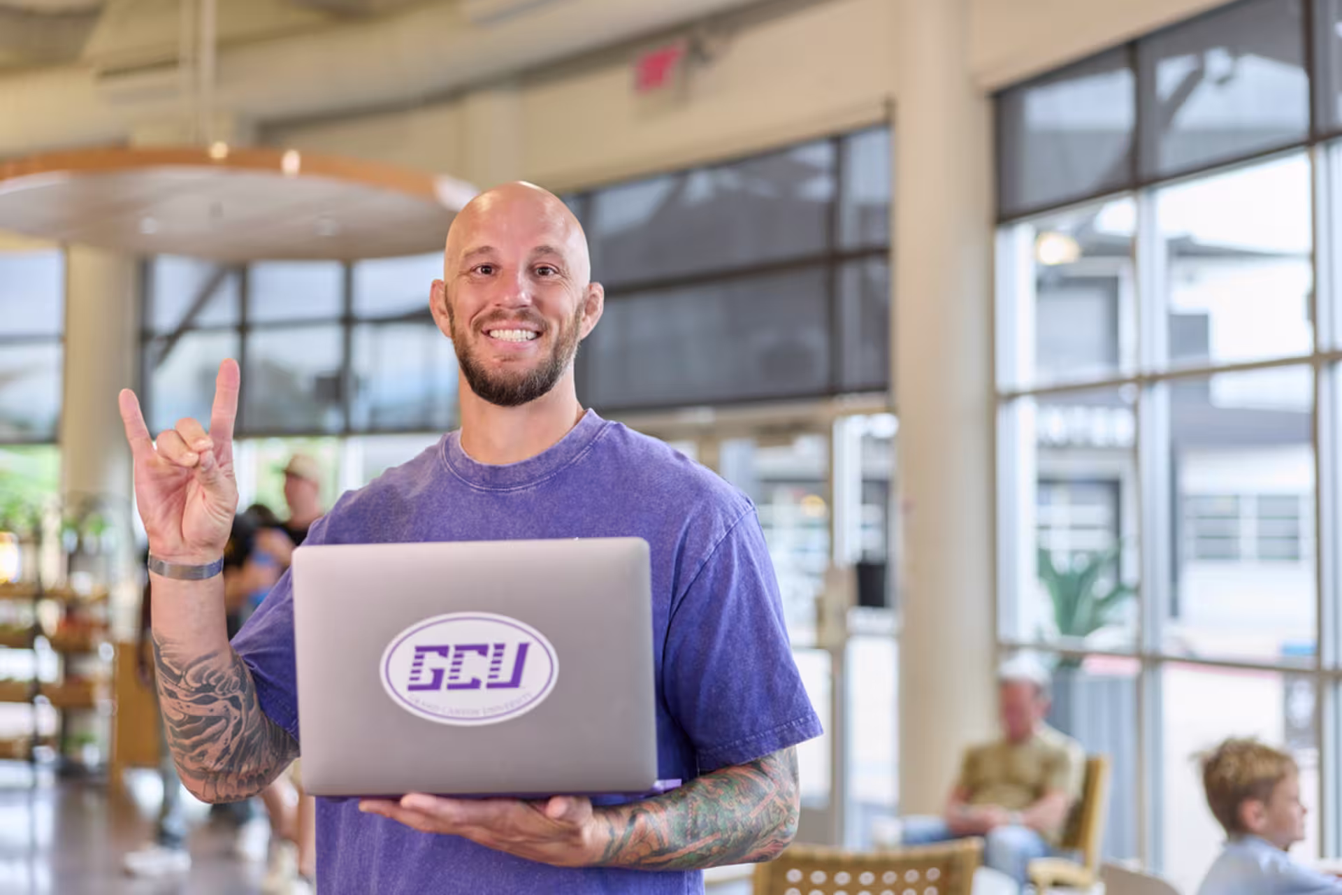 Male bachelor's student in a purple t-shirt standing on campus while holding a GCU laptop.