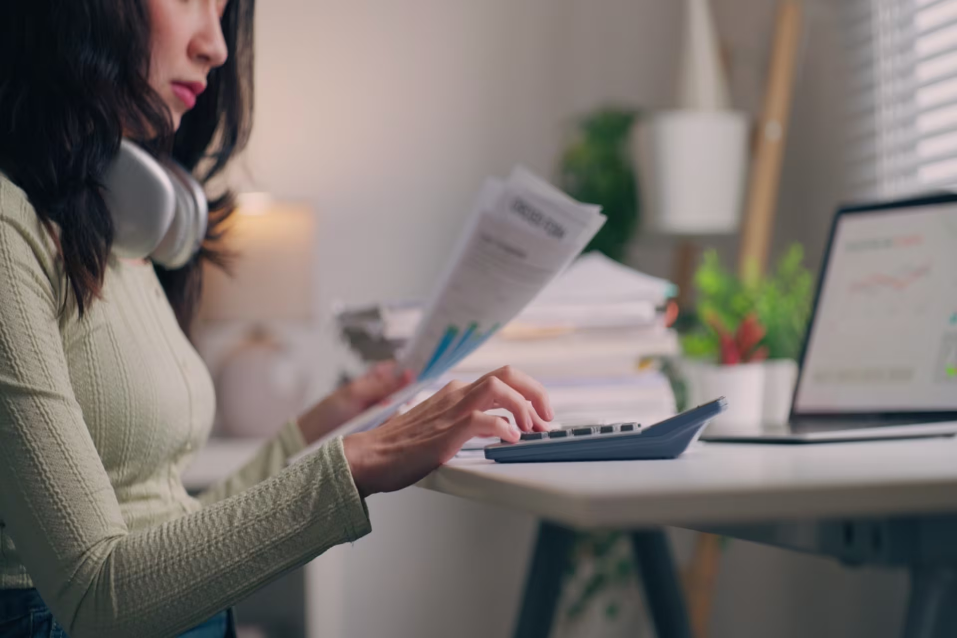 A female accountant reviews financial documents and uses a calculator at a home office desk.