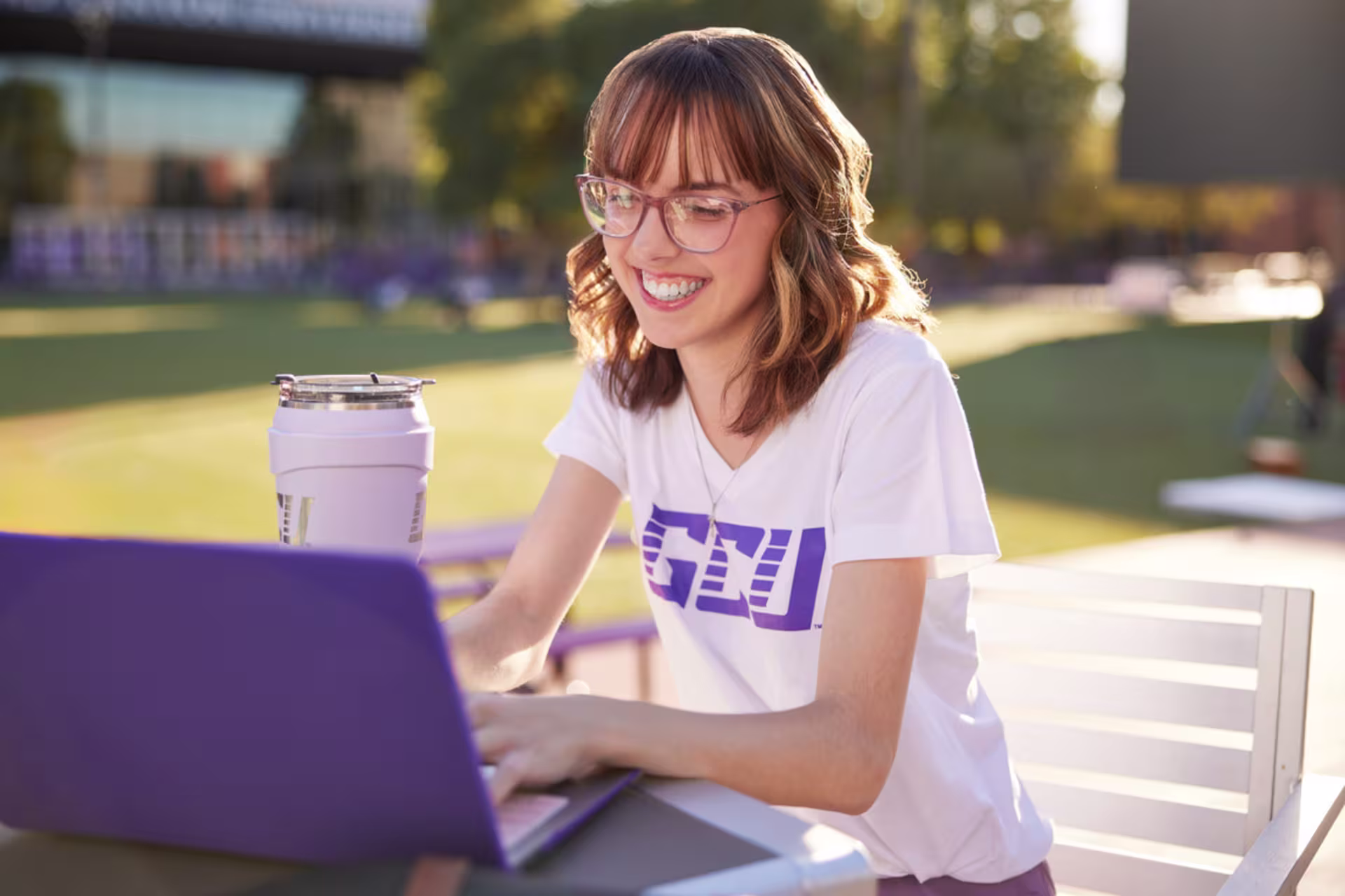 Smiling student wearing a GCU T-shirt, working on a laptop outdoors at a campus table. 