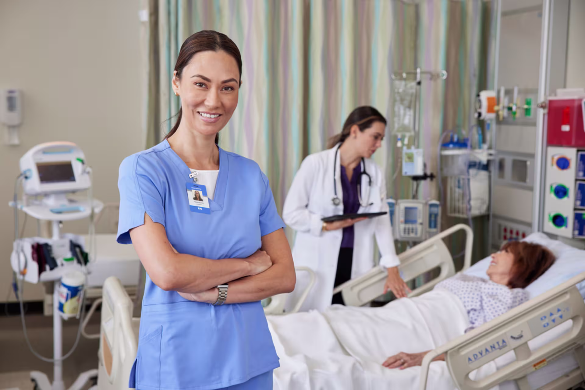 Registered nurse stands smiling in a hospital room while a physician cares for a patient in a hospital bed in the background.