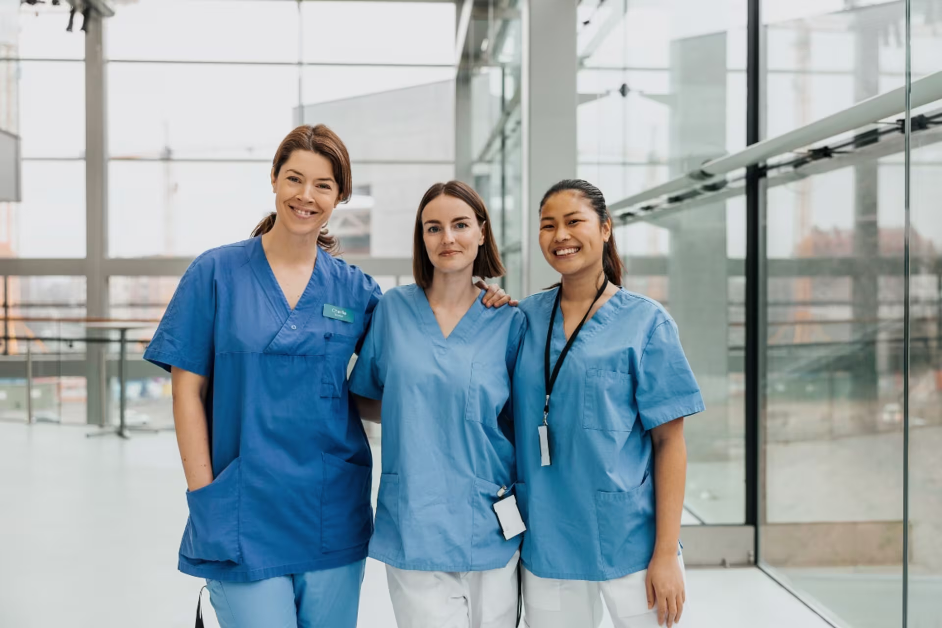 Three female nursing MSN student in scrubs standing together in a medical facility smiling at the camera.