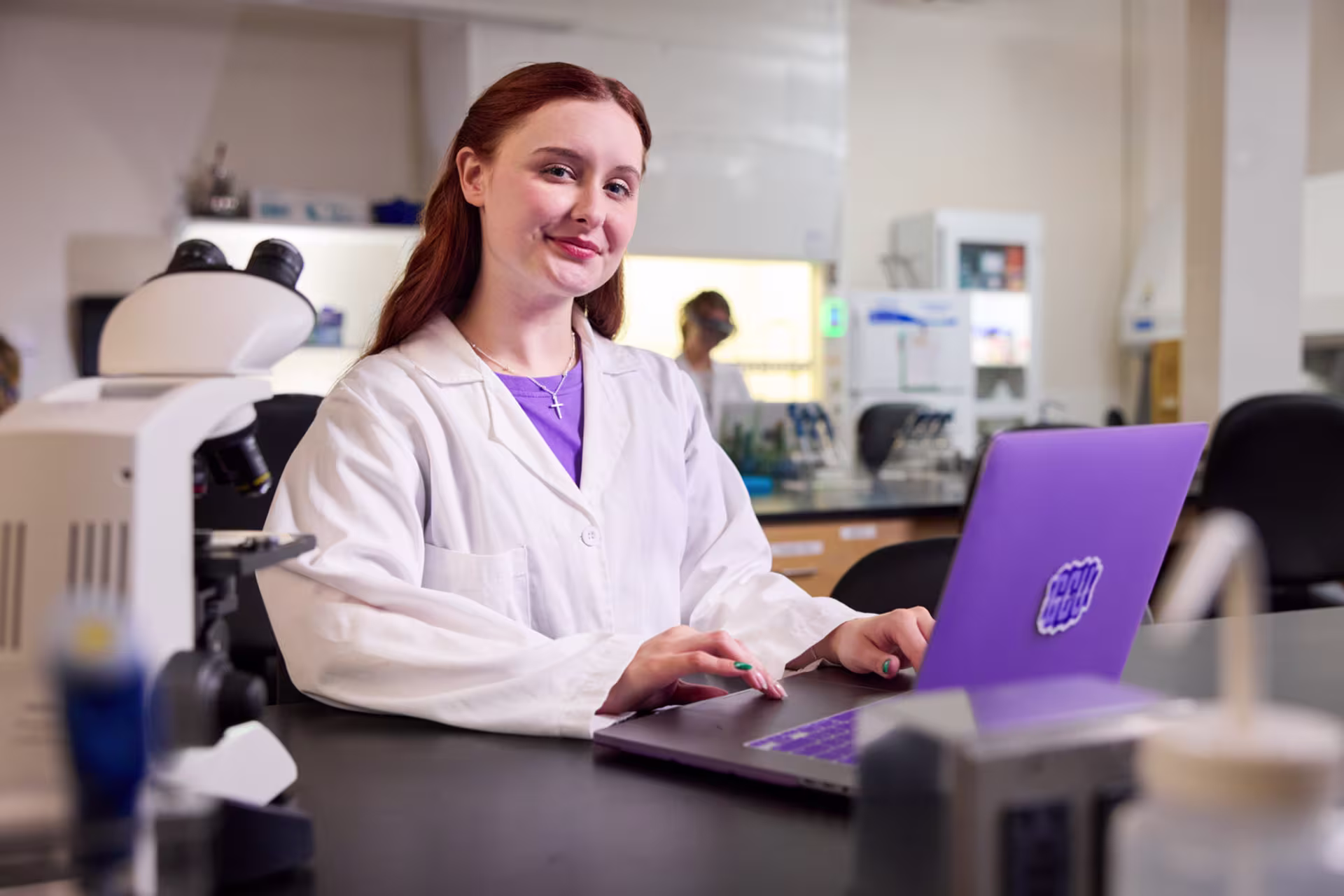 Science student in a white lab coat working on a laptop beside a microscope in a laboratory.