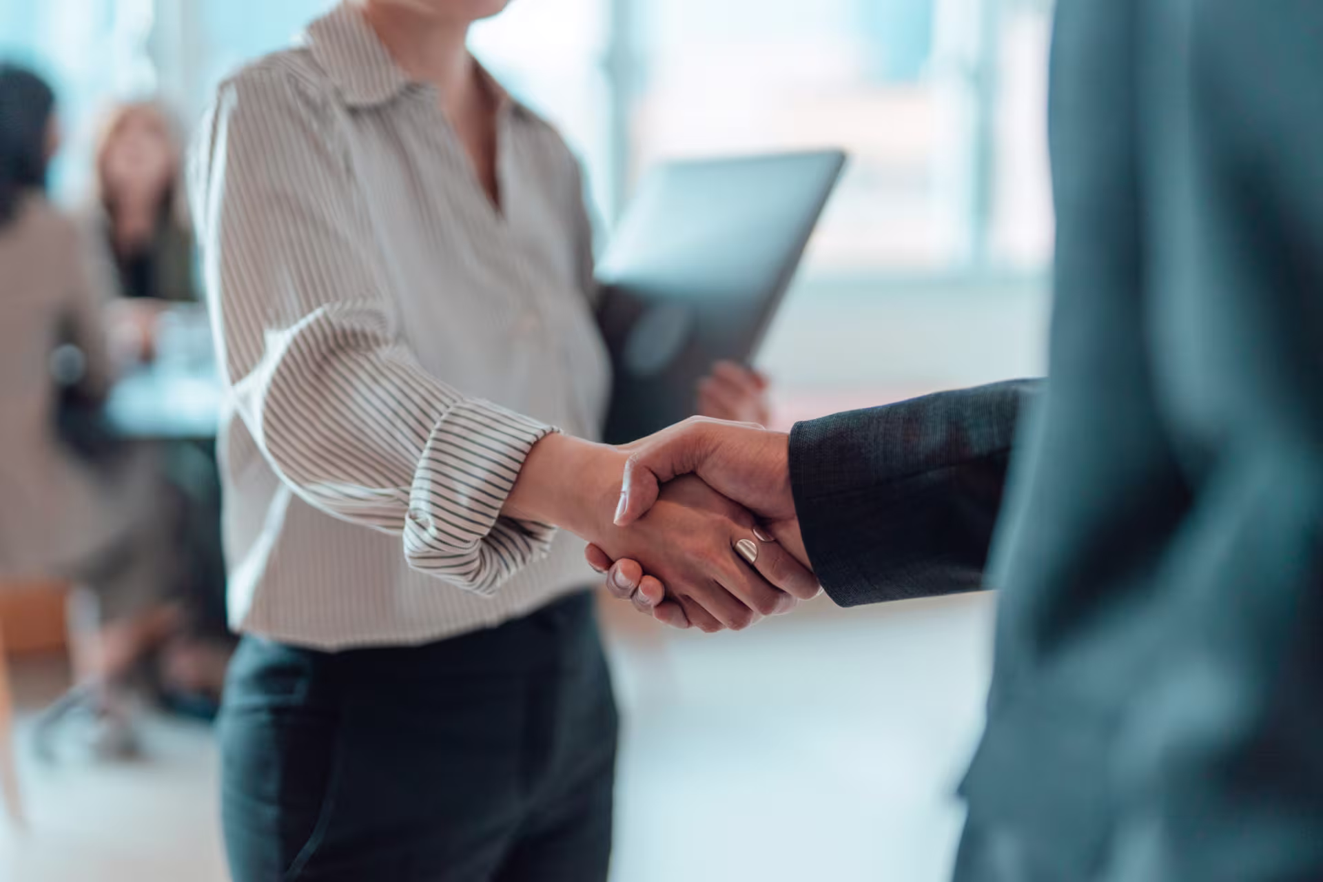 Business professionals shake hands in a modern office setting, with one person holding a laptop and colleagues blurred in the background.