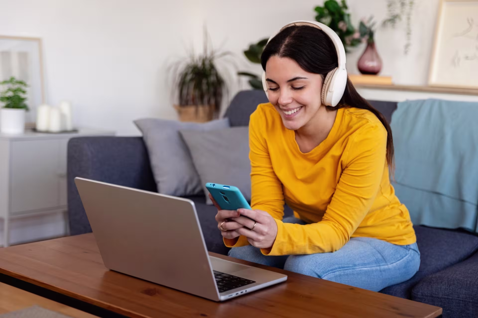A female social media student wearing headphones uses a smartphone while studying with a laptop.