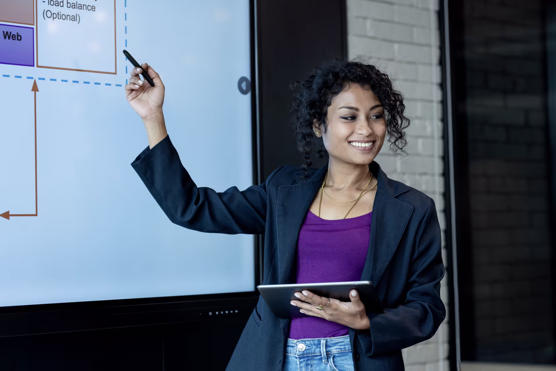 A person holding a tablet points to a digital presentation screen displaying a workflow diagram during a business meeting.
