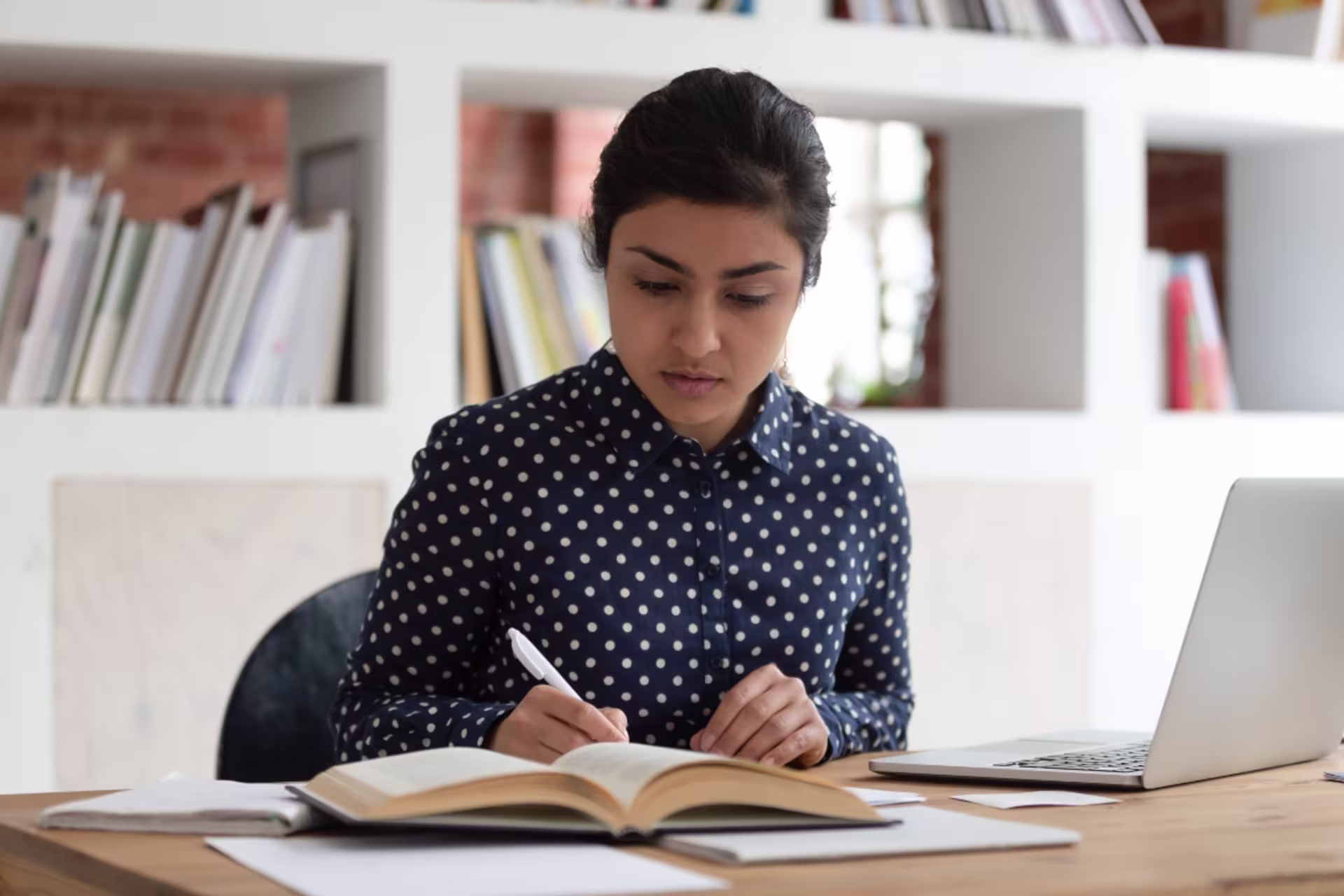 A certificate student takes notes while studying from a textbook and laptop.