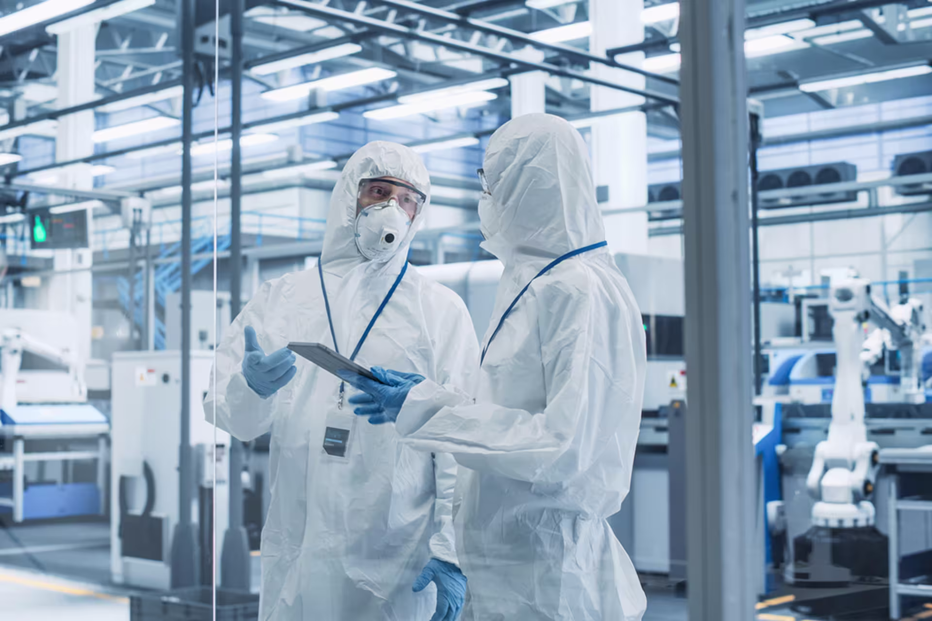 Assembly test technicians in a factory setting in white clean suits discuss plans on a tablet.
