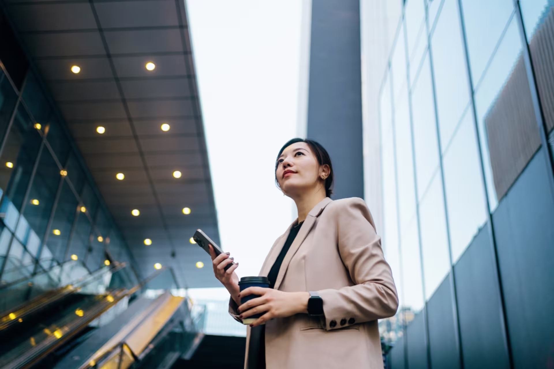 Confident doctoral graduate holding a smartphone outside a modern glass office building, looking upward with an ambitious and professional demeanor.