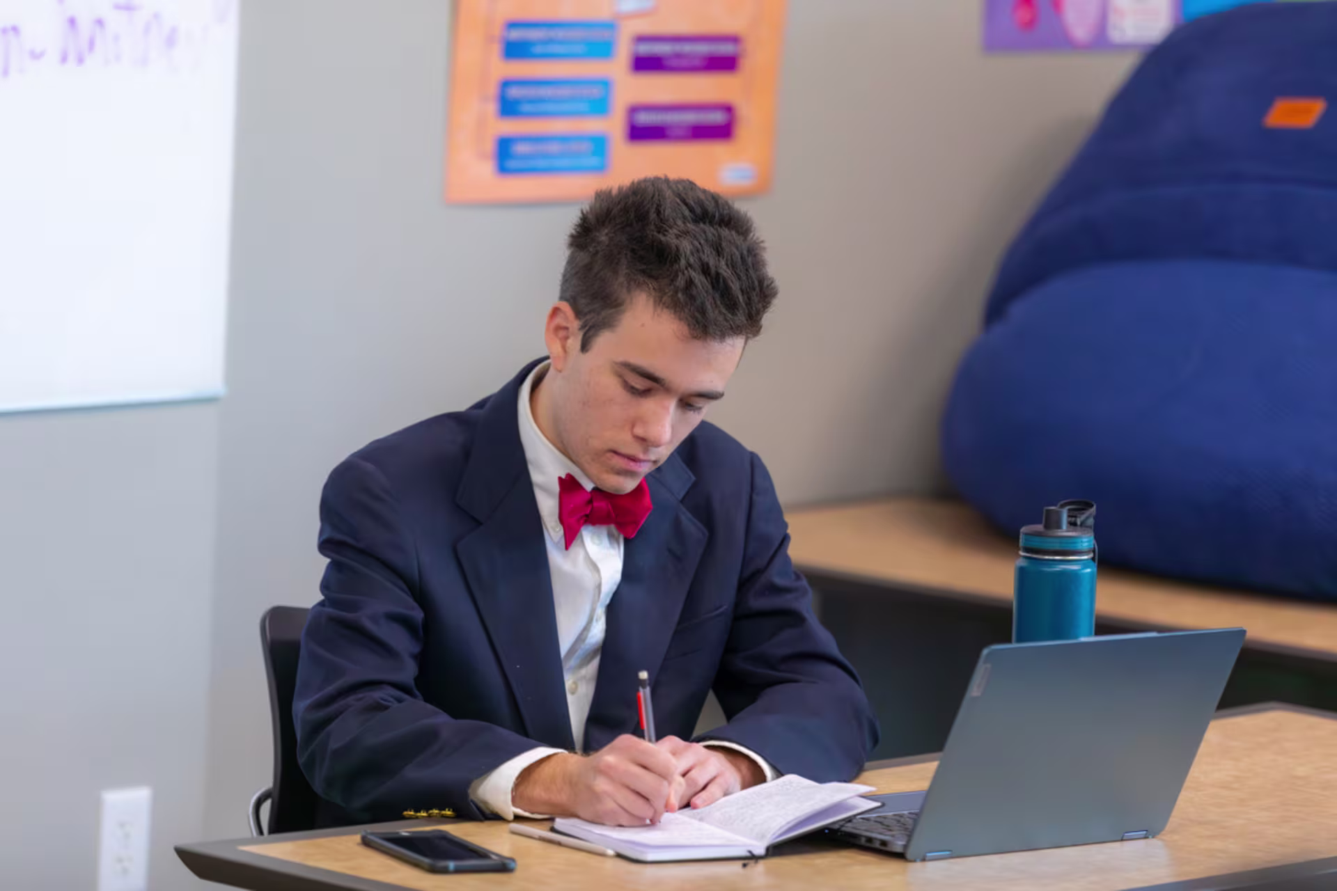 GCU PhD student in a suit and red bow tie taking notes at a desk with a laptop and water bottle, focused in a classroom or study environment.