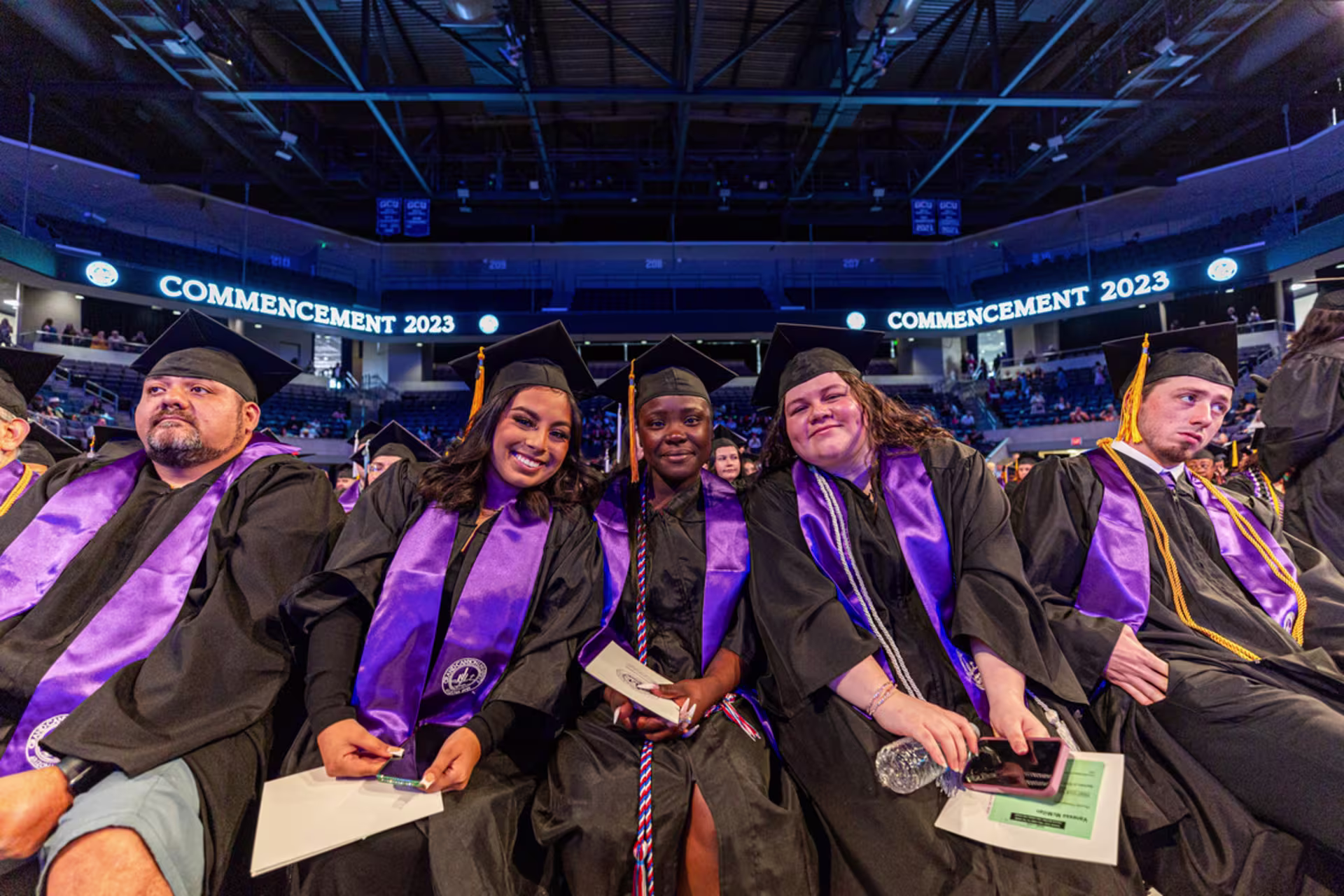 GCU doctoral graduates wearing black caps and gowns with purple stoles seated together at a commencement ceremony.