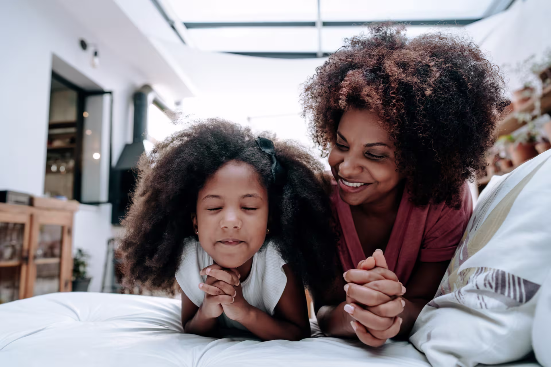 Person and child kneeling on a couch at home with hands clasped, appearing to pray together in a bright living room.