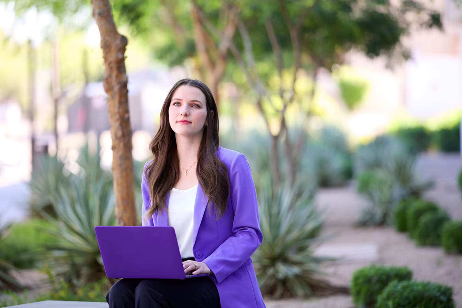 Woman in a purple blazer using a laptop while seated outdoors in a landscaped setting.