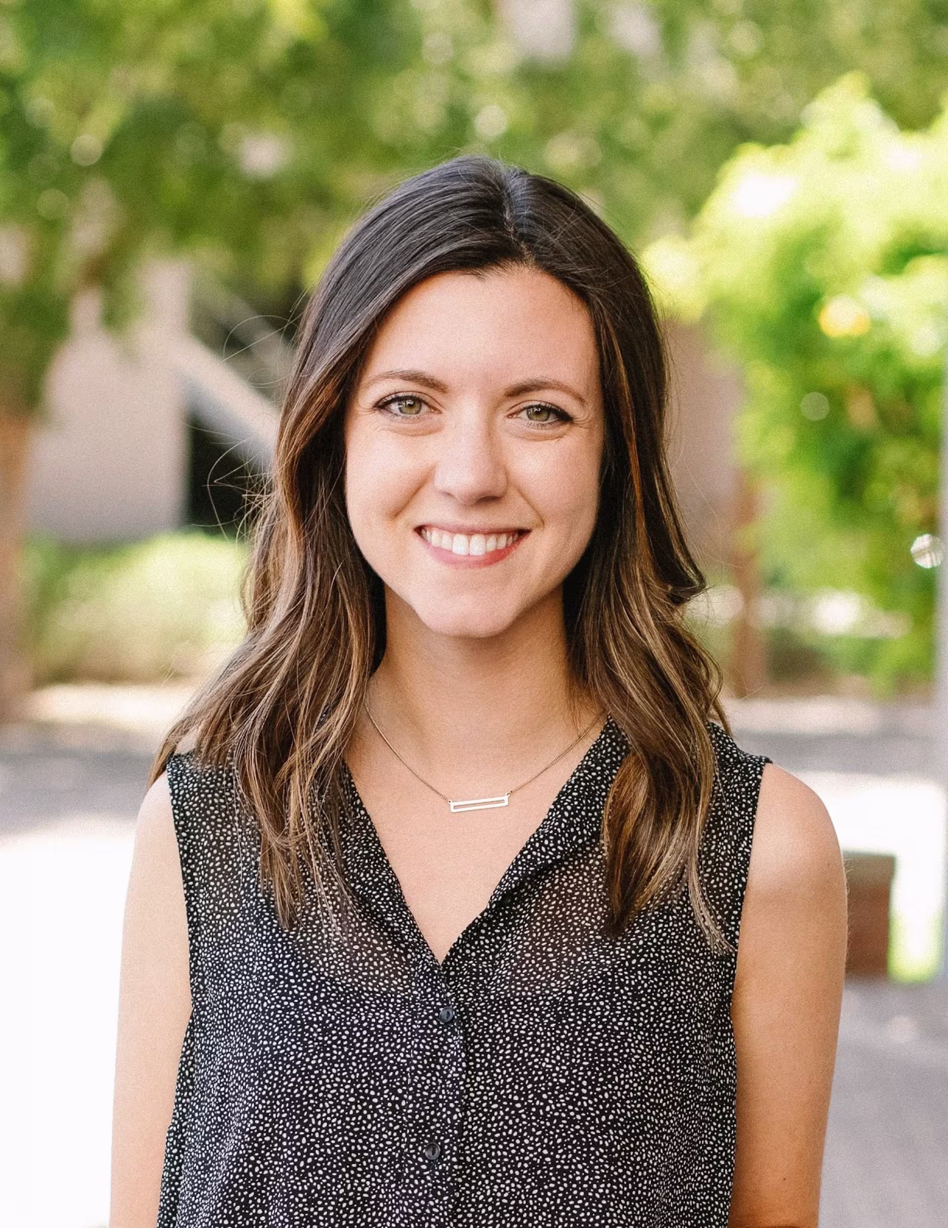 Professional outdoor headshot of a person with shoulder-length dark hair wearing a black patterned sleeveless blouse, standing in front of a softly blurred green background.