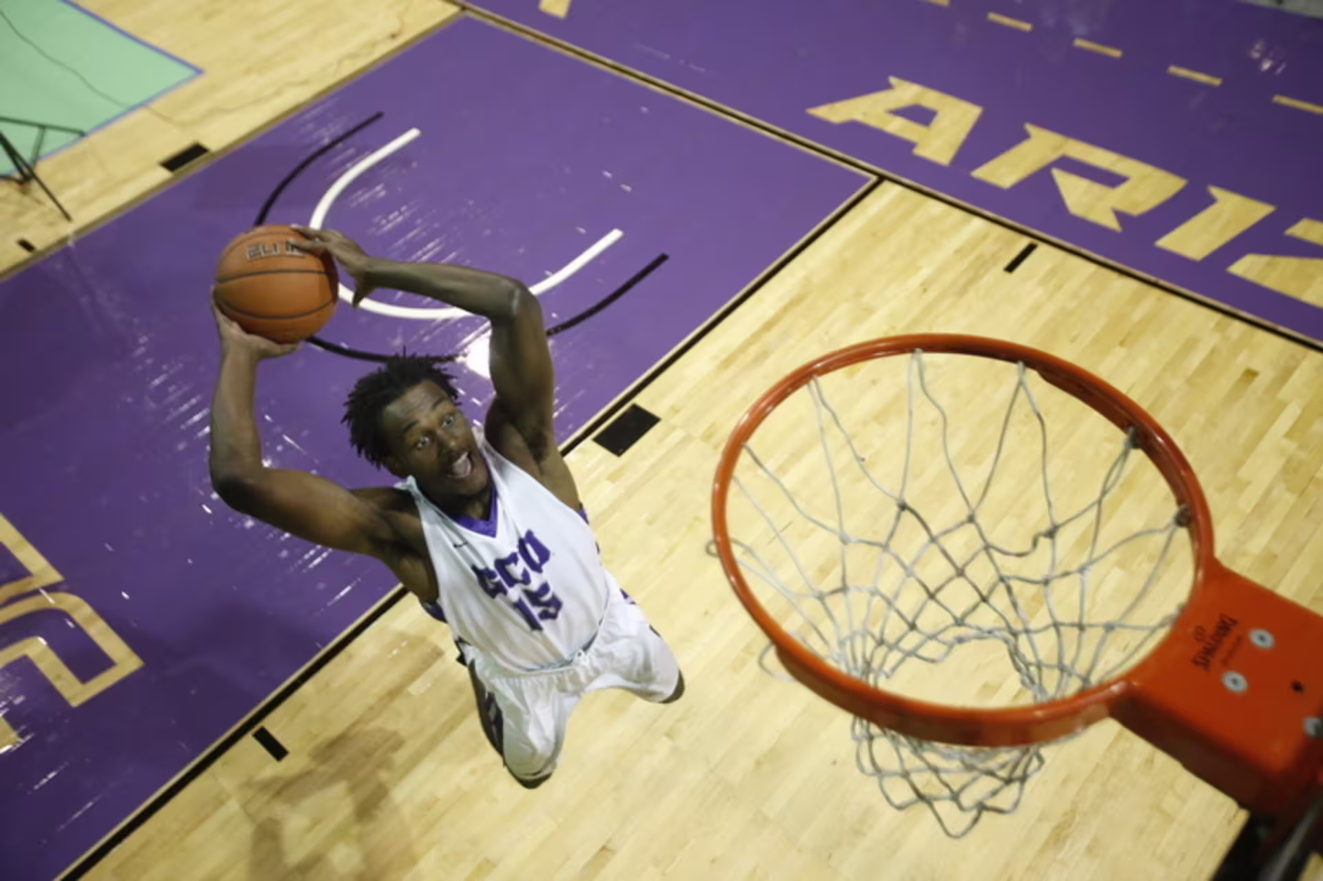 Basketball player in a white jersey leaps toward the hoop for a dunk, viewed from above on a hardwood court with purple painted lanes and arena lettering.