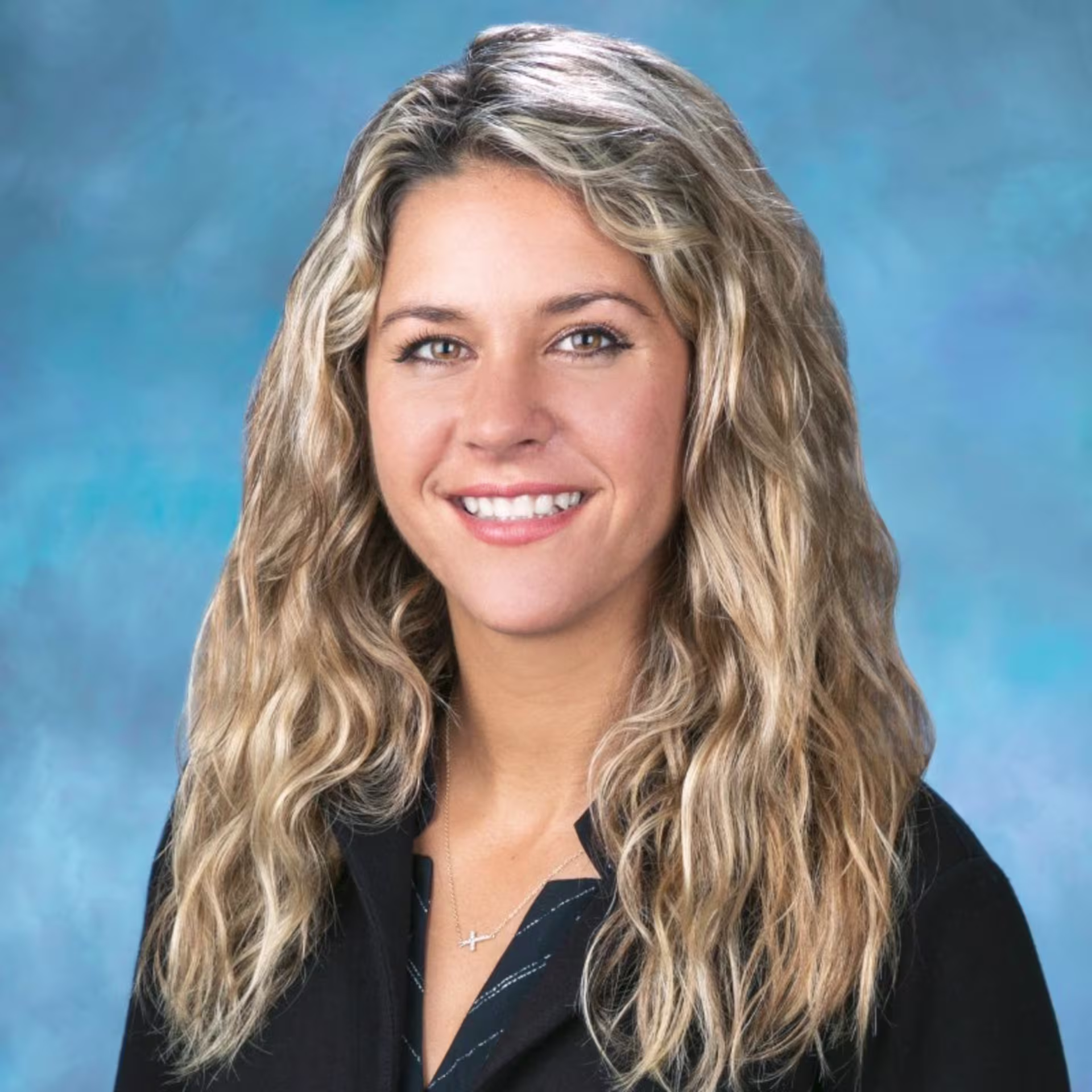Professional headshot of a person with long wavy hair wearing a dark blazer and necklace, photographed against a blue studio background.